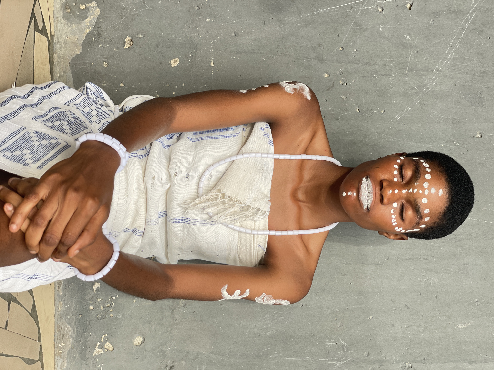 A person lies on a gray concrete surface, presumably a floor or ground. The person is of African descent, with dark skin and short black hair. White markings are painted on their face, primarily around the eyes and on the lips. They are wrapped in a white cloth with blue stripes. White bracelets are on both wrists, and a white beaded necklace is around their neck. There are white markings painted on their arms as well. The person appears to be lying down with their eyes closed or partially closed. The setting appears to be outdoors, as the surface shows signs of wear and weathering. The image was taken in Badagry, Nigeria.