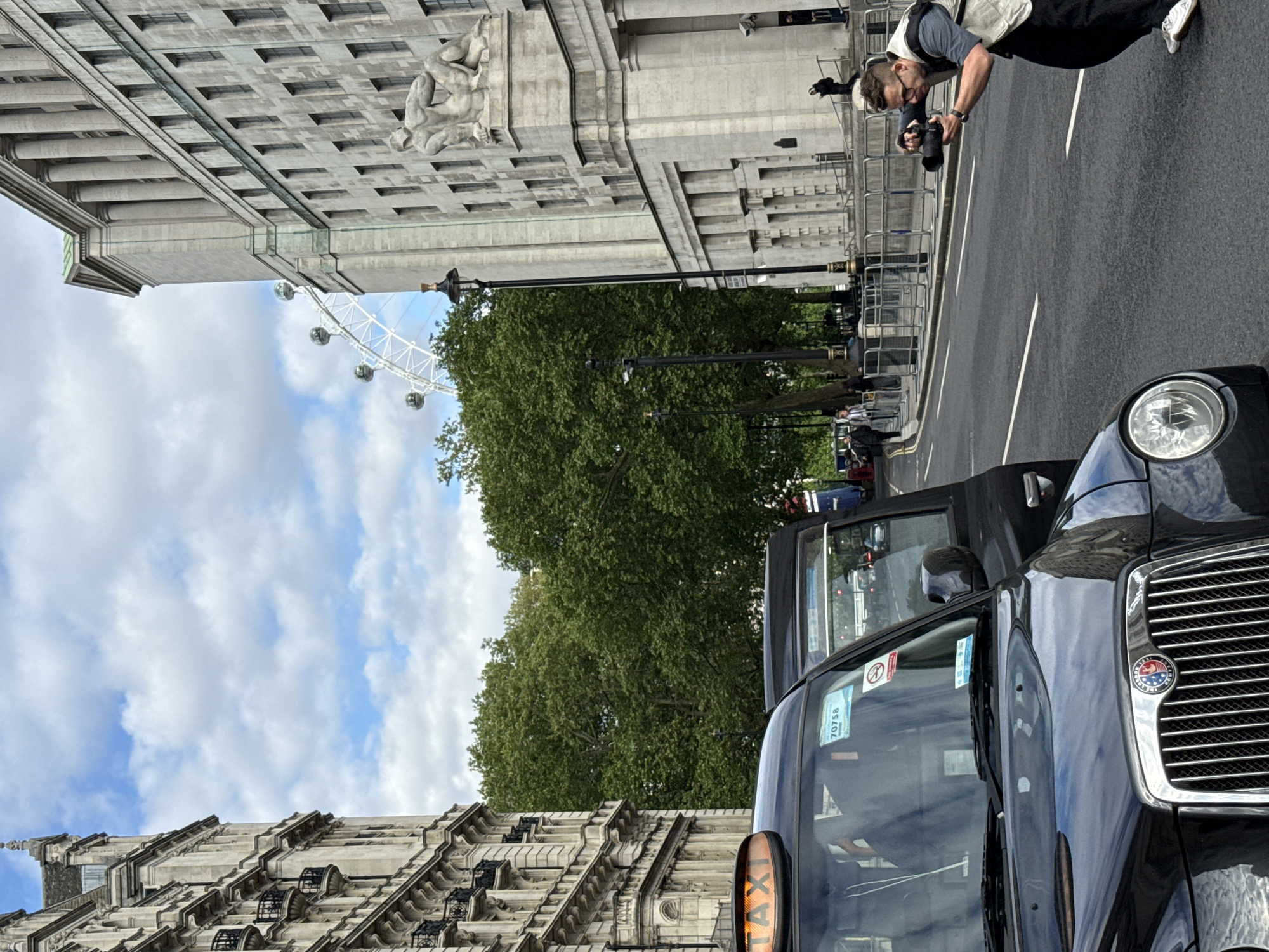 The image shows a street scene in London, United Kingdom, featuring a black London taxi in the foreground. In the background, partially obscured by trees, is the London Eye. A photographer is seen taking a picture, standing on the road next to a metal barrier. The buildings on either side of the street have ornate architectural details. The sky is partly cloudy.