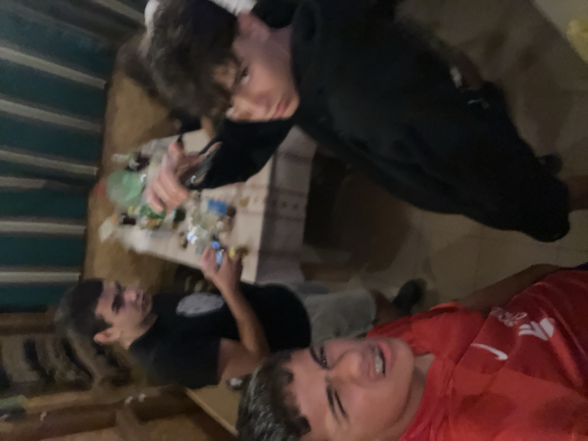 Three young men are depicted in a dimly lit indoor setting. The man in the foreground on the left wears a red t-shirt and makes a grimacing facial expression. To his right, a second man in a black hoodie looks away from the camera, his right arm extended. In the background, a third man in a black t-shirt and light-colored shorts holds a phone. A table in the background is cluttered with bottles and other items. The overall environment appears to be a casual gathering, possibly in a home or a recreational space.
