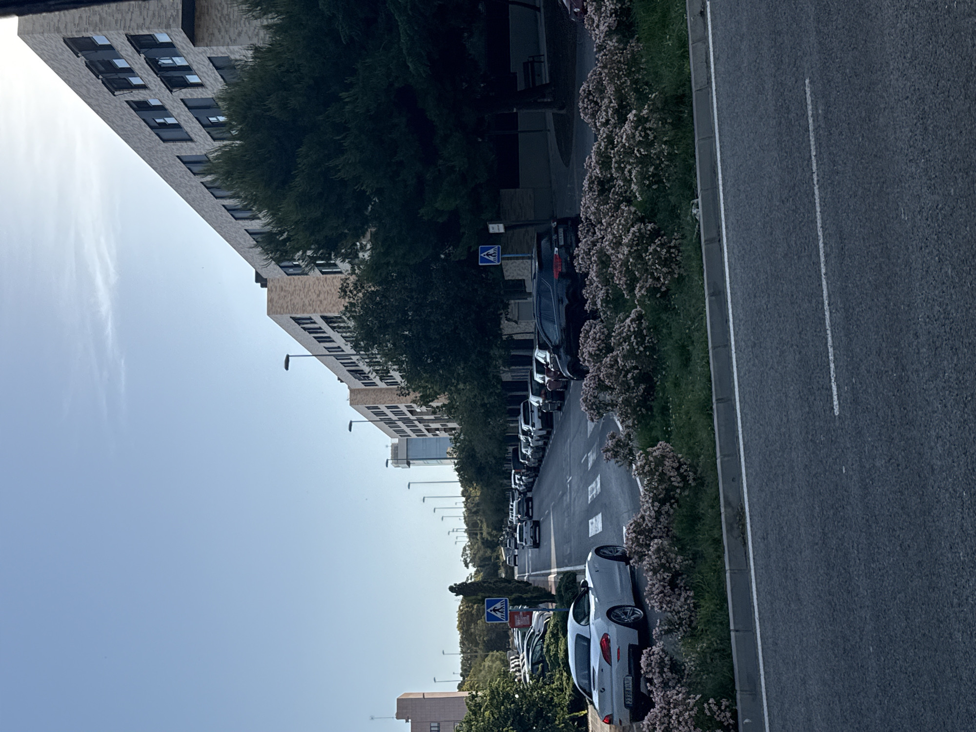 The image shows a street scene in a modern urban environment, likely a city. The street is lined with parked cars on both sides, and there is a white BMW Z4 in the foreground on the left, parked facing left. A pedestrian crossing is visible in the center of the image. On the right side of the street, a multi-story building with a modern architectural design, featuring light-colored brickwork and numerous windows, stretches into the distance. There are also trees and streetlights along the street. The sky is clear and blue, suggesting daylight. The overall impression is one of a well-maintained, contemporary urban area.
