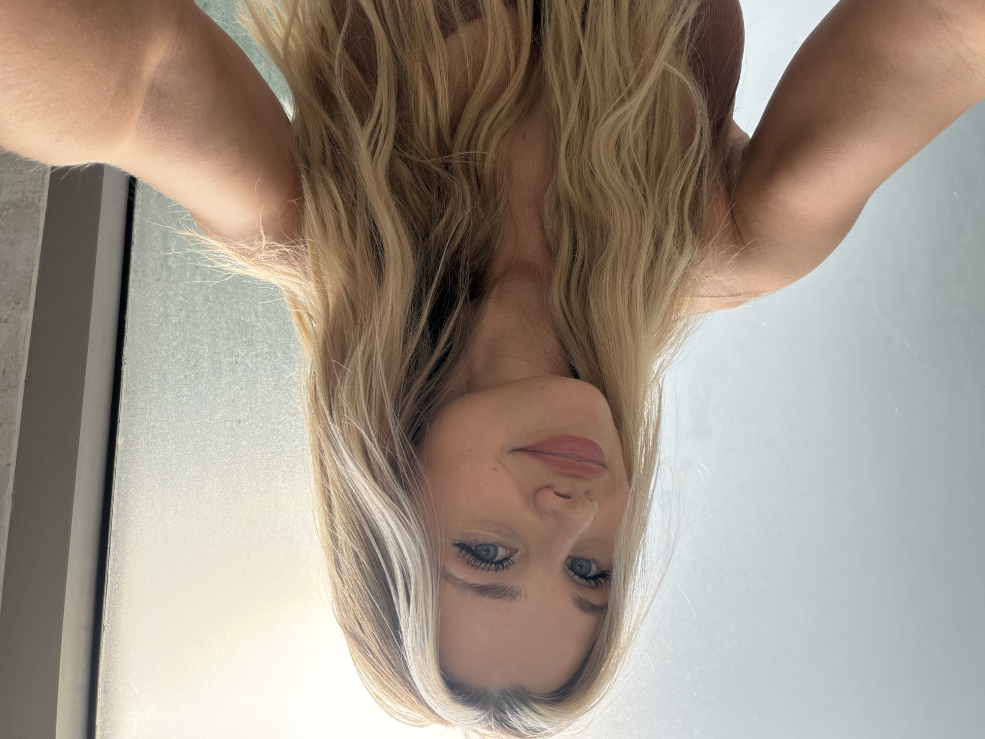 The image shows a young woman with long, blonde, wavy hair. She is wearing a light brown tank top and has blue eyes and fair skin. She is smiling and looking at the camera, with her right arm extended as if taking a selfie. The background appears to be a wall with frosted glass and a vertical dark strip, possibly a doorway or window frame. The lighting is bright, coming from the right side of the image, creating a soft glow. There are no discernible features that would indicate the location is in Thep Krasatti, Thailand.