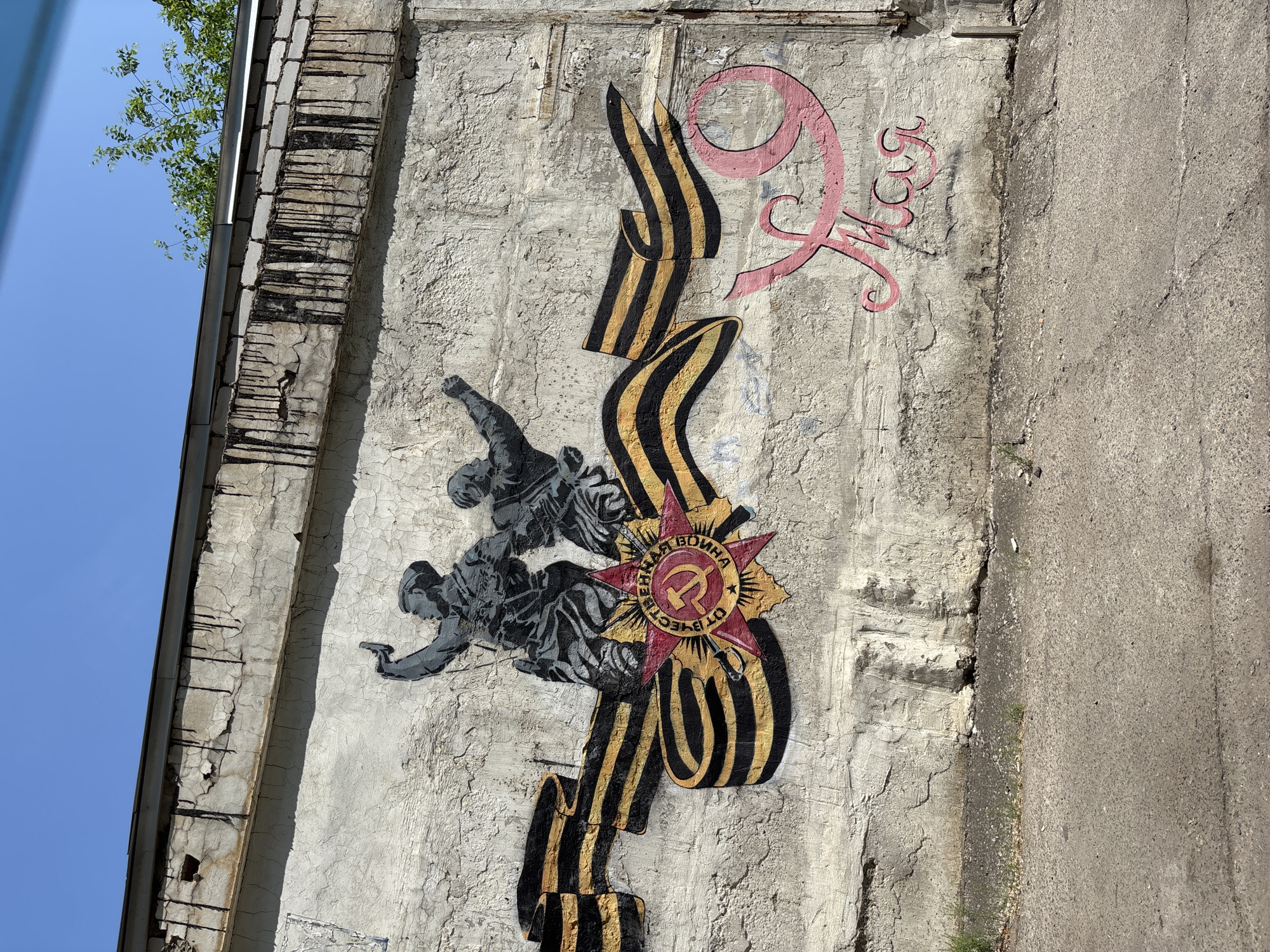 The image shows a mural on a concrete wall in Blagoveshchensk, Russia. The mural depicts two figures in military uniforms raising their arms in victory, with a St. George's ribbon and a Soviet-era medal. The figures are rendered in a stencil style with black and grey tones. To the right of the mural, the word "May" is written in pink Cyrillic script. The wall itself is weathered and cracked, with evidence of water damage and peeling paint along the top edge. The ground in front of the wall is asphalt with some patches of grass growing through cracks. The sky above is clear blue. The mural is a commemoration of Victory Day, celebrated on May 9th in Russia.