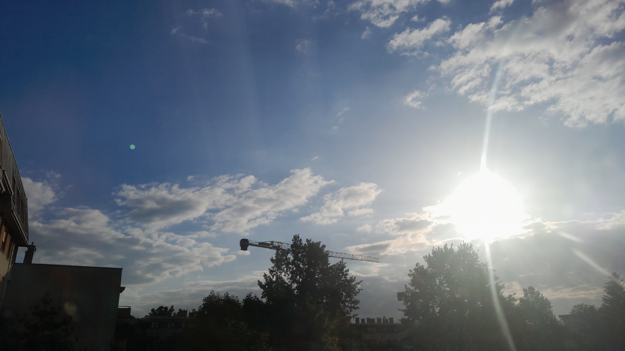 The image captures a cityscape under a bright, sunlit sky. The sun, positioned towards the right side of the frame, is a brilliant, almost blinding source of light, with rays emanating outwards and piercing through scattered clouds. A tall construction crane is visible in the mid-ground, silhouetted against the sky and partially obscured by trees. In the foreground, on the left, is the side of a building, appearing to be an apartment complex with visible balconies and window structures. The overall scene suggests a developing urban environment bathed in the strong light of either sunrise or sunset. The green orb in the sky is likely a lens flare.