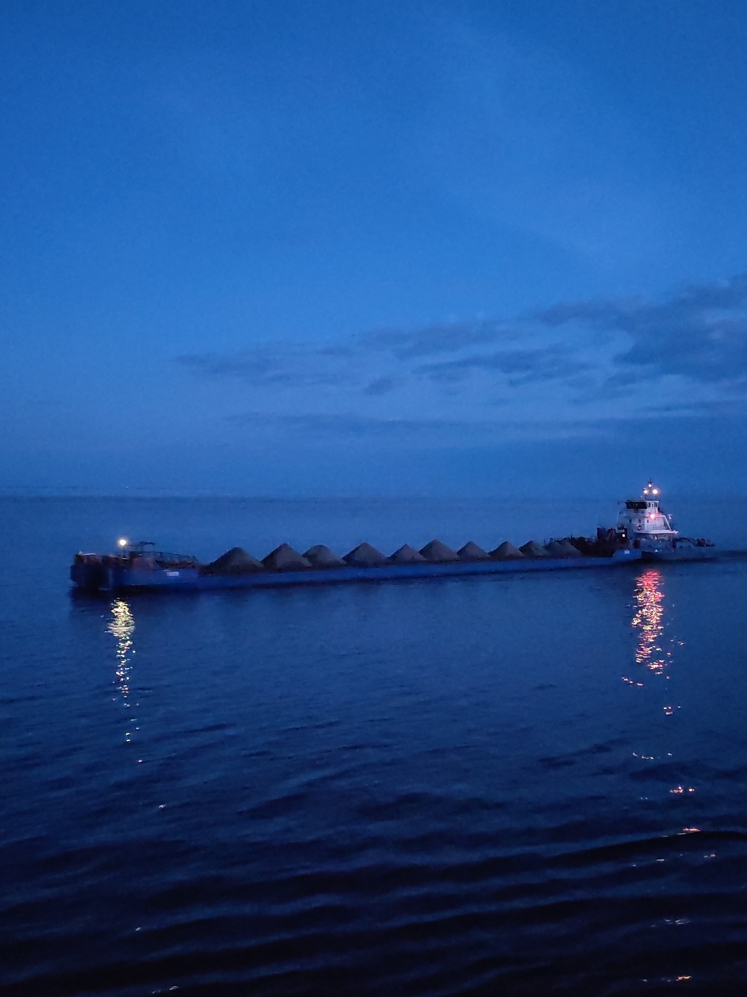 A wide shot depicts a barge traveling on a body of water, likely at dusk or during the night. The barge is long and loaded with several cone-shaped piles of material, potentially sand or gravel. A tugboat is visible at the front of the barge. Reflections of the barge's lights are visible on the water surface. The sky is a dark blue with some clouds visible along the horizon. The city of Legkovo, Russia is not clearly discernible from this image.