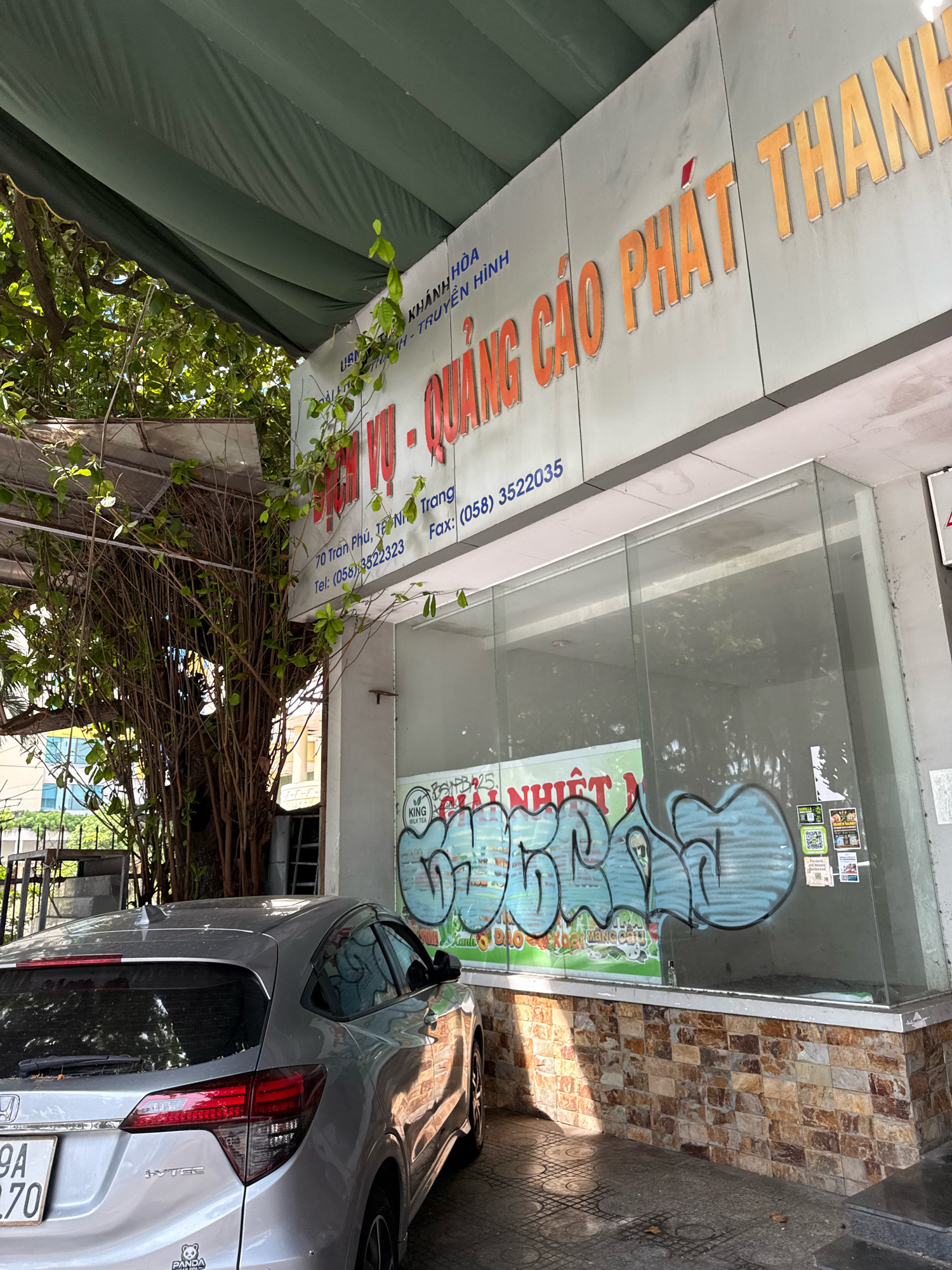 A silver Honda HR-V is parked in front of a commercial establishment in Nha Trang, Vietnam. The establishment features a large sign with orange lettering that reads "DỊCH VỤ - QUẢNG CÁO PHÁT THÀNH". Below this, smaller text indicates an address "70 Trần Phú, TP. Nha Trang" and contact information, "Tel: (058) 3522323 Fax: (058) 3522035". The building has large glass windows, one of which is covered with graffiti in light blue and black spray paint. A portion of a tree with many branches and leaves is visible on the left side of the image, partially obscuring the building. The ground is paved with patterned concrete tiles. The license plate of the car is visible and reads "9A 370.70".
