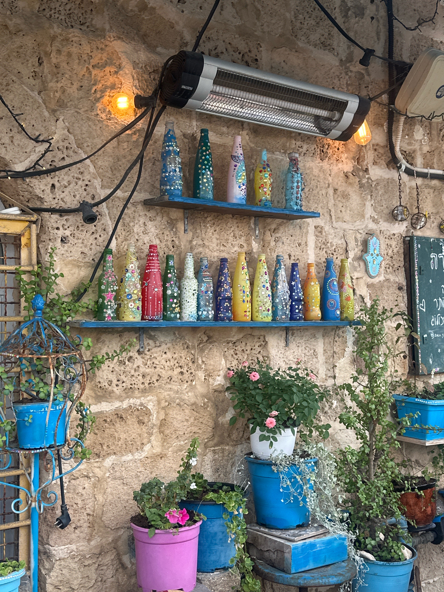 The image depicts an outdoor scene in Tel Aviv, Israel, likely a cafe or shop exterior, with a rough stone wall as the backdrop. Two blue shelves are mounted on the wall, adorned with an assortment of colorful, decorated glass bottles. The bottles are painted and embellished with beads, glitter, and other decorative elements, arranged in a whimsical display.
Above the shelves, a silver electric heater is mounted on the wall, with a visible heating element. A bare incandescent light bulb hangs nearby, casting a warm glow. Various black electrical wires and cords are visible, connecting the heater and light fixtures.
To the left of the shelves, a vintage-style blue metal birdcage holds a potted plant. Adjacent to it, a yellow-framed window pane with a lattice design is visible.
Below the shelves, several potted plants add greenery to the scene. A large pink pot contains a flowering plant with pink blossoms. To its right, a vibrant blue pot holds more greenery. Further to the right, a smaller brown pot also contains a plant. Blue boxes and a blue stool are stacked beneath some of the plants, adding to the eclectic decor.
A dark green chalkboard with white lettering, possibly writing in Hebrew or Arabic, is mounted on the wall to the right. A small blue Hamsa hand decoration is also attached to the wall.
The overall impression is of a charming, bohemian-style outdoor space, characteristic of some of Tel Aviv's older neighborhoods like Jaffa or Neve Tzedek, known for their artistic ambiance and unique boutiques. No people are visible in the image.