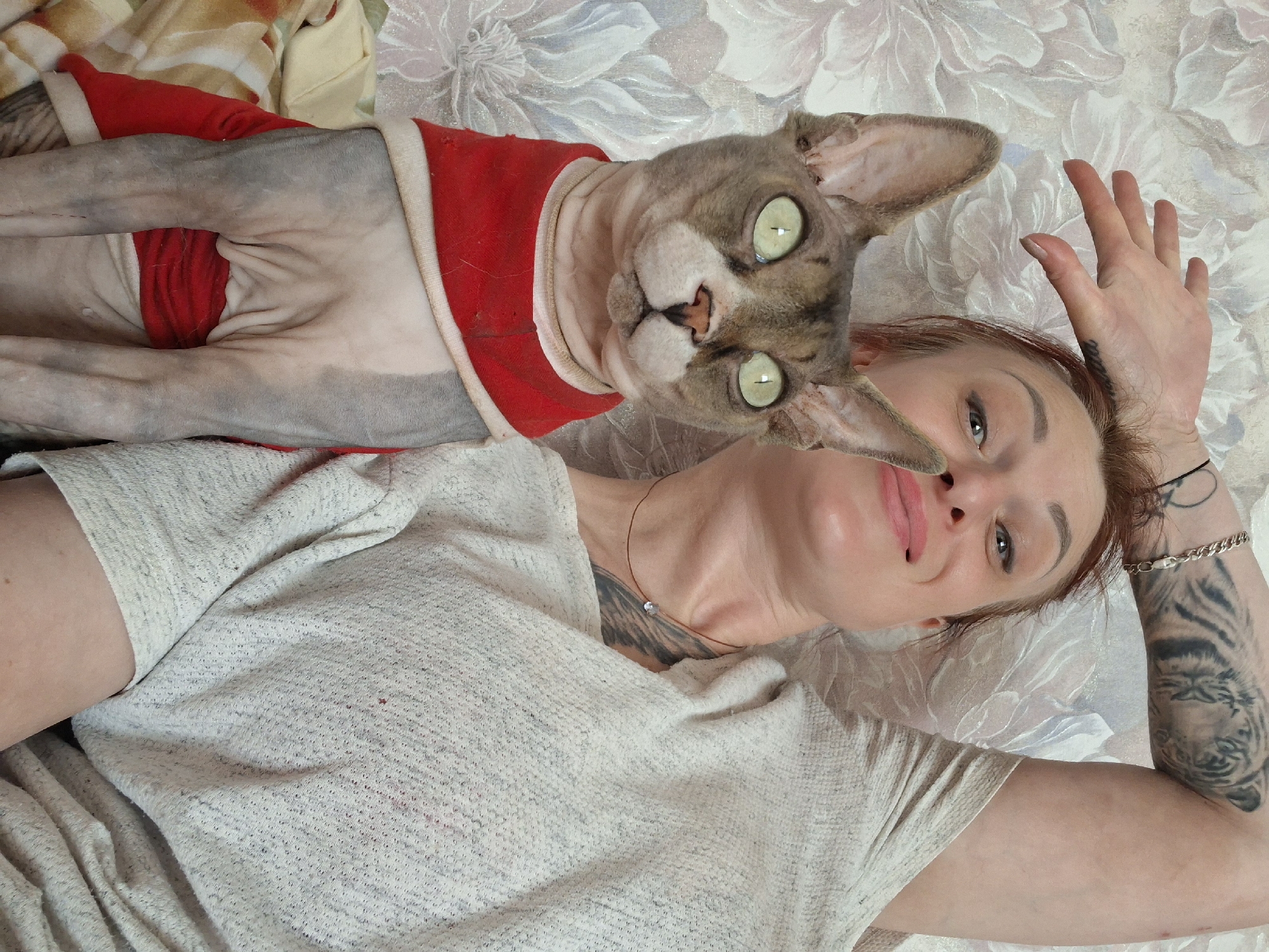 A woman with red hair and tattoos is lying on a bed next to a Sphynx cat wearing a red garment. The cat is in the foreground, looking directly at the camera with wide green eyes. The woman is in the background, propped up on an elbow with her hand near her face. The bed has a floral-patterned comforter and a textured blanket. The scene appears to be indoors, likely in a bedroom. The location in Забалоцкі сельскі Савет, Belarus cannot be determined from the image.