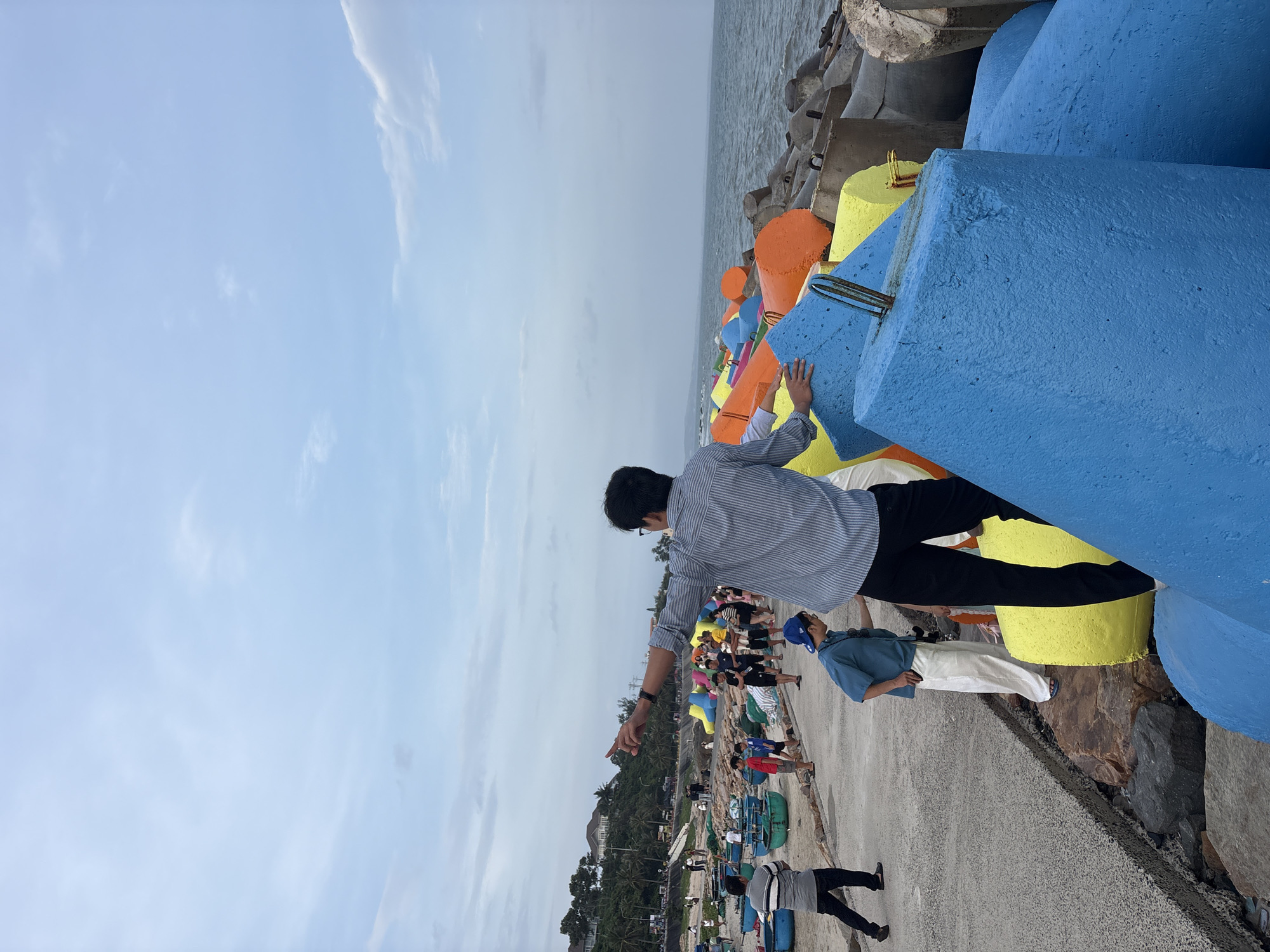 The image captures a scene at a coastal area in an unknown city in Vietnam. In the foreground, a man in a striped shirt points towards the ocean, with his back to the camera. He is standing on a large, bright blue concrete barrier. Behind him, a group of people are gathered near the water, some walking along a concrete path, others observing the surroundings. Several colorful, conical concrete structures painted in blue, yellow, and orange are visible along the coastline, interspersed with more traditional grey concrete breakwaters. In the background, traditional Vietnamese fishing boats are lined up on the shore, and lush green hills dotted with buildings rise above the coastline. The sky is overcast with scattered clouds.