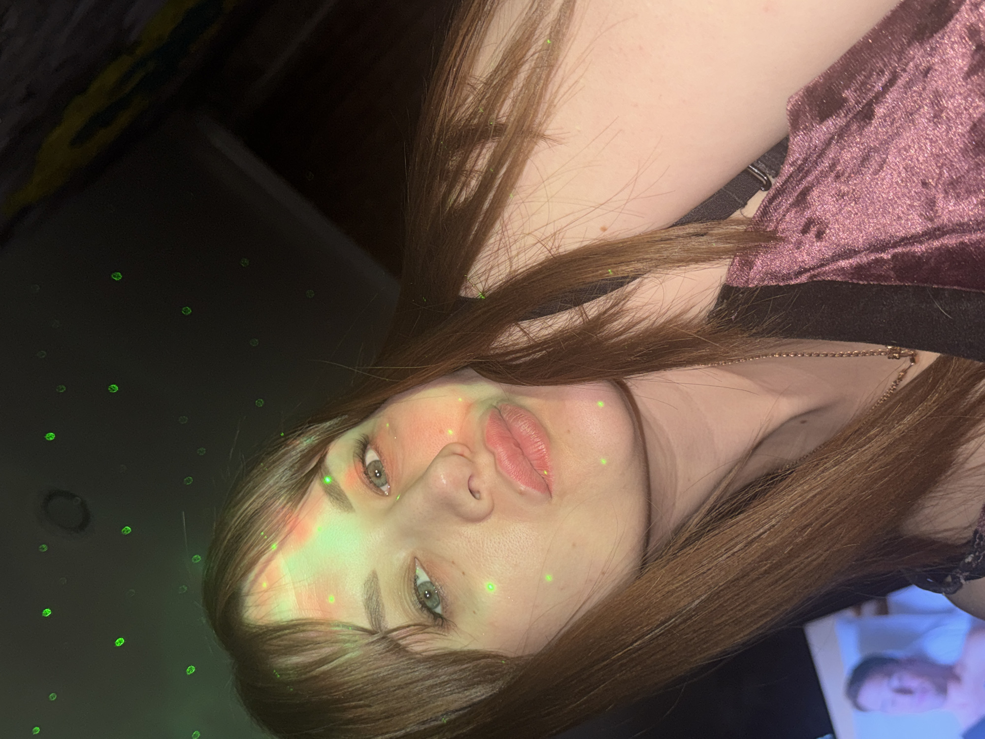 A woman with light skin, green eyes, and light brown hair is facing the camera, turned slightly to her right. She has make-up on, including blush and lip gloss. She wears a purple, velvet-like garment with a black top or strap visible underneath. A thin, gold-colored necklace is visible around her neck. Her face is illuminated by a green light that also casts several small, green dots onto the dark background. The location is unknown and there is no evidence to suggest it is in the city of Chaikovsky, Russia. A screen displaying images of people is visible in the bottom right.