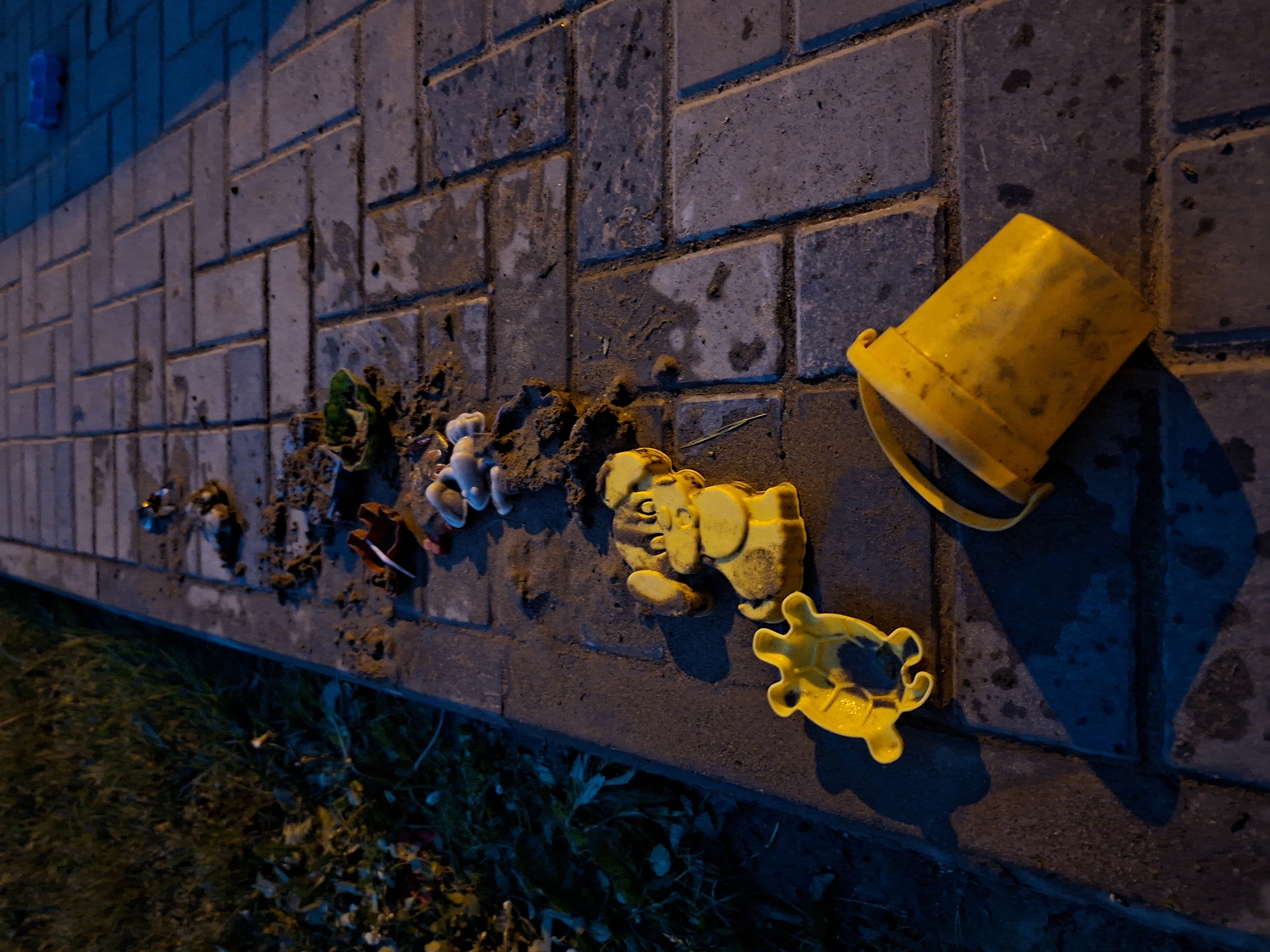 The image depicts a sidewalk in Votkinsk, Russia, at ground level. The sidewalk is paved with interlocking gray bricks, some of which are wet. To the left of the sidewalk, a patch of dark green grass and fallen leaves is visible. Scattered along the edge of the sidewalk and on the bricks are children's toys, predominantly yellow plastic sand molds in the shapes of a dog, a turtle, and a bucket. Other toys, including a red and white plastic toy and some smaller, indistinct plastic figures, are also present amidst piles of dirt and sand. The scene is illuminated by artificial light, casting distinct shadows.