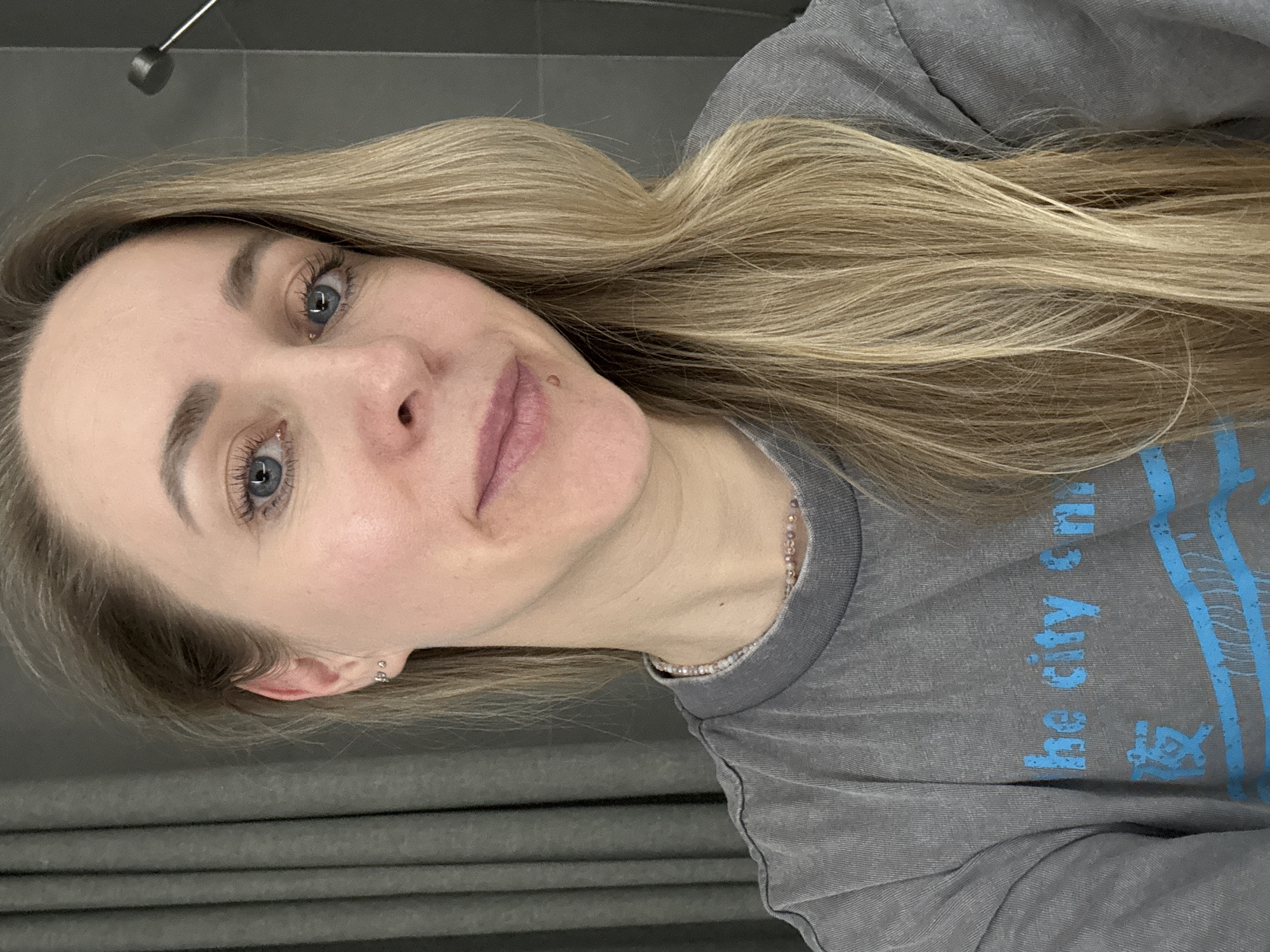 A young woman with long, wavy blonde hair and blue eyes is featured in a self-portrait, smiling gently at the camera. She appears to be in her 20s or early 30s and has fair skin, with a small mole visible above her upper lip on the right side. She is wearing a light grey, possibly stone-washed, t-shirt with light blue text printed on it. Partially visible text on the shirt reads "the city en" and another line below it. A delicate, light-colored beaded necklace is visible around her neck, and a small, sparkly hoop earring adorns her left ear.
The setting appears to be indoors, with a plain, dark grey wall behind her. On the left side of the frame, a darker grey texture suggests a curtain or blinds. A dark, cylindrical object on a rod hangs from the ceiling in the upper background. The lighting is soft and even, suggesting an indoor environment without strong direct sunlight. The woman's expression is calm and pleasant, indicating a relaxed atmosphere. The image is taken in a selfie style, with the woman looking directly into the lens. The location context is Chaikovsky, Russia.
