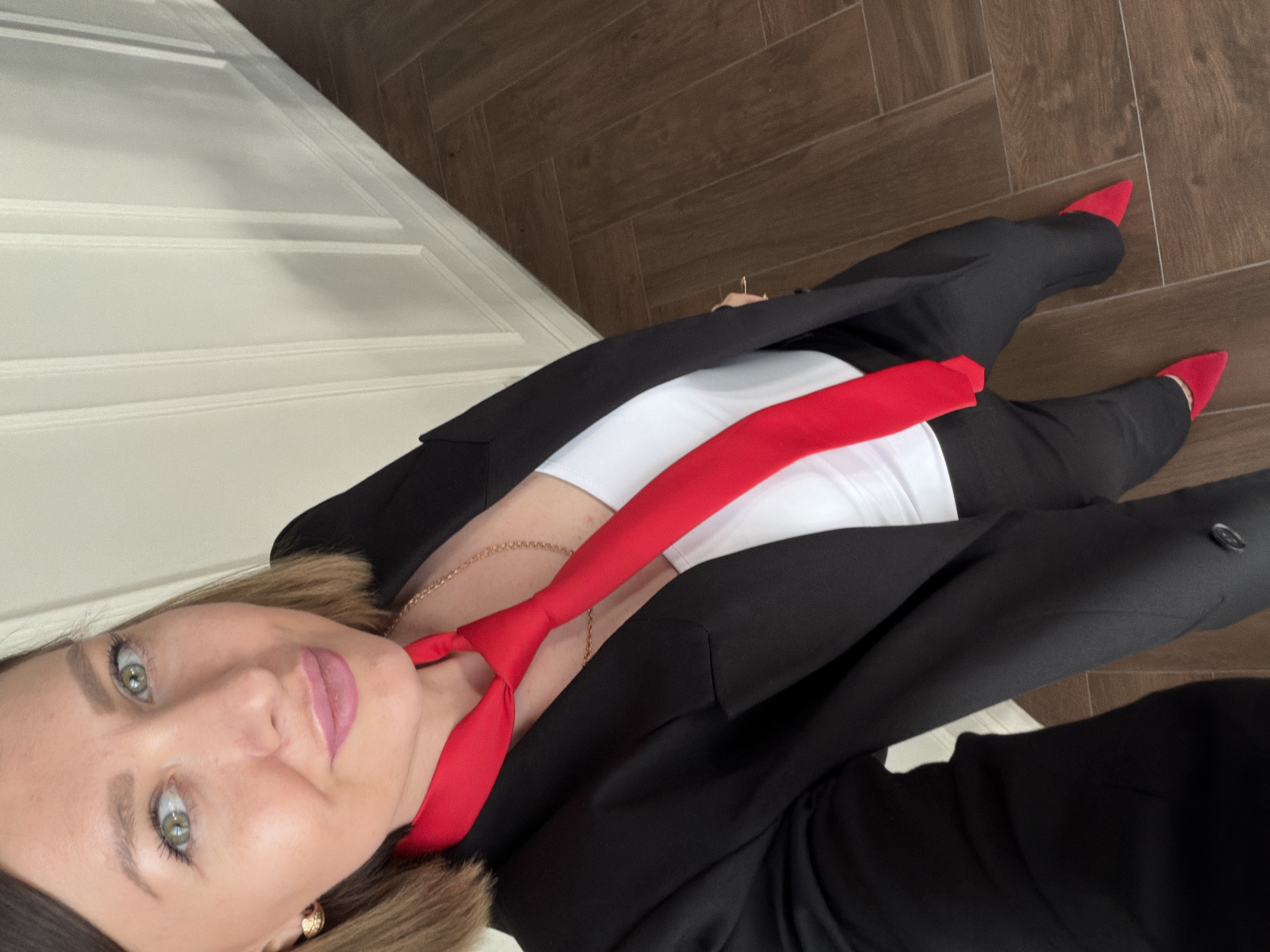 A woman with shoulder-length brown hair, wearing a black blazer, white top, and a red necktie stands in a room with brown tile flooring. She is wearing red pointed-toe shoes and gold earrings. A white paneled door is visible on the left. The location is not possible to discern as Rostov-on-Don, Russia.