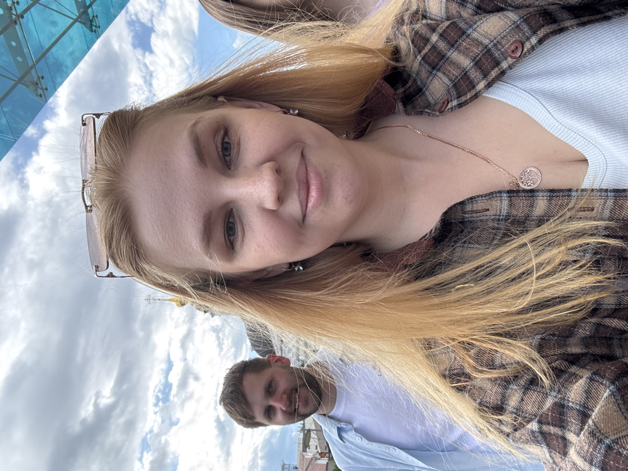 A selfie of a fair-skinned woman with long blonde hair, wearing a brown plaid shirt over a light blue top and a rose gold necklace with a round pendant. She is smiling at the camera. In the background, slightly out of focus, a man with a beard is also smiling. The background also shows a cloudy sky, part of a blue glass structure, and the rooftops of buildings. Based on the spired architectural style visible, the location is likely in Russia, specifically Rusanovo. The subjects are interacting by posing for a selfie together.