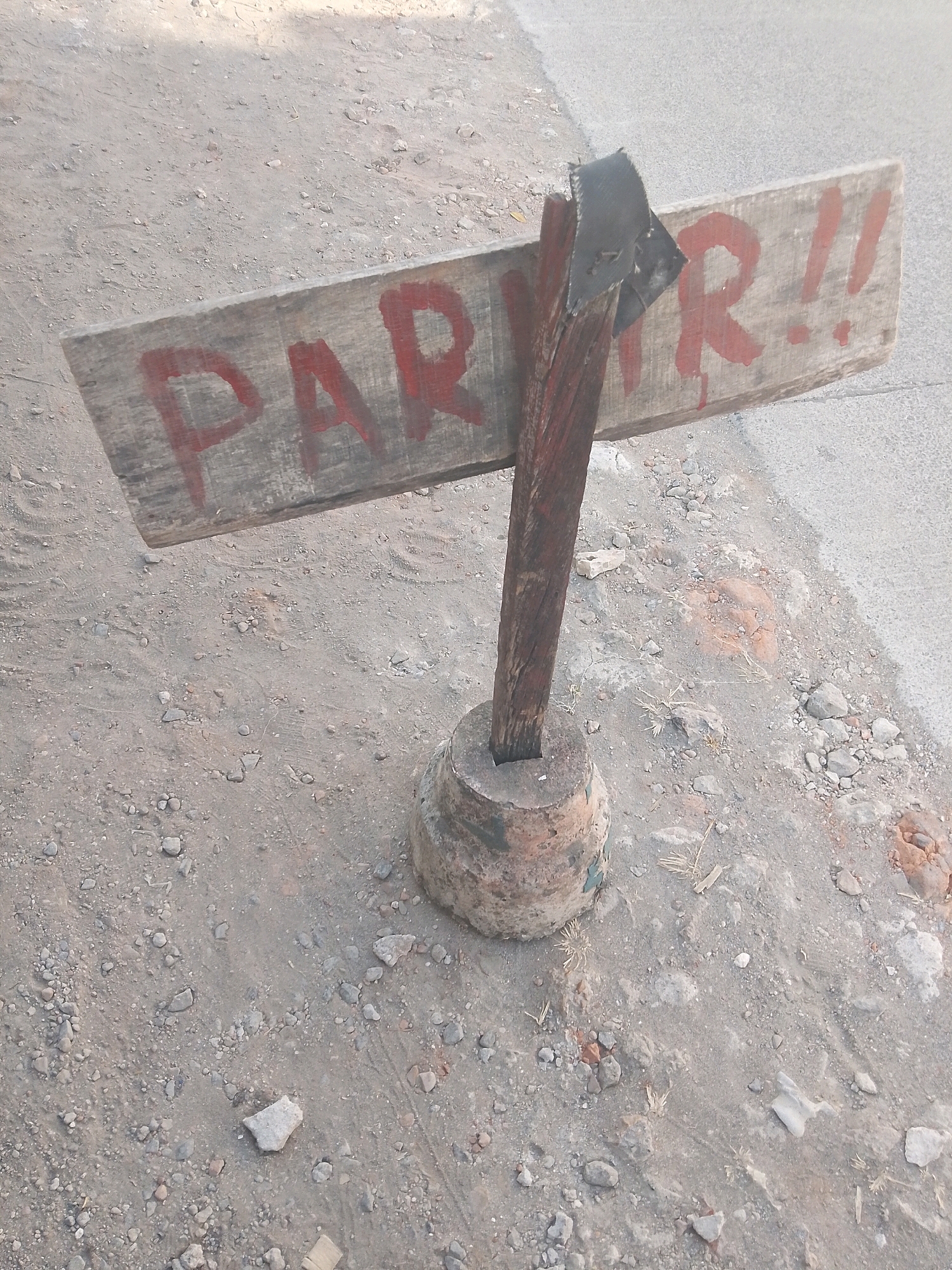 A weathered wooden sign with red lettering that reads "PARR!!" stands tilted on a rustic base of concrete and a wooden post. The sign is attached to the post with what appears to be black tape. The surrounding environment is a dusty, gravelly ground with scattered small rocks and debris. The scene is outdoors, likely on a street or pathway, given the presence of asphalt or a similar paved surface in the background. There are no people visible in the image. The overall impression is one of a makeshift, possibly temporary, sign in a somewhat unkempt outdoor area.