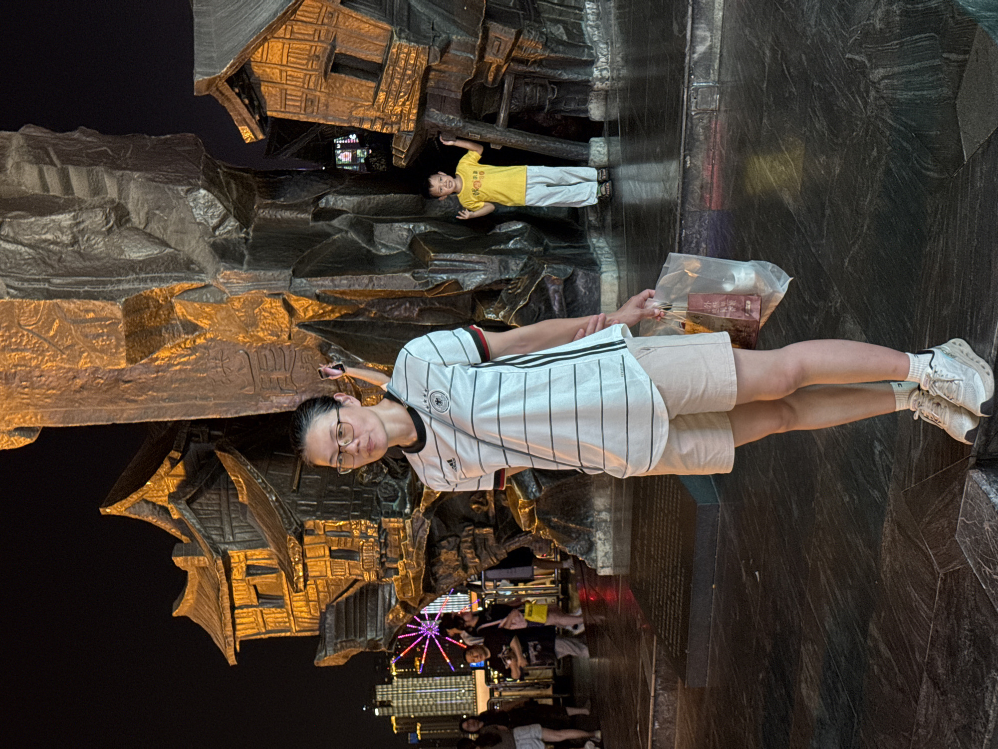 In Yuzhong District, China, at night, a woman wearing glasses, a white and black striped athletic shirt, and light-colored shorts stands in the foreground. She is holding a clear plastic bag containing items. Behind her, a young boy in a yellow t-shirt and light pants stands, making a peace sign with his fingers. The background features a large, dark, abstract sculpture with gold-colored architectural elements that resemble traditional Chinese houses. In the distance, city lights and a Ferris wheel are visible. The ground is paved with dark, reflective tiles.