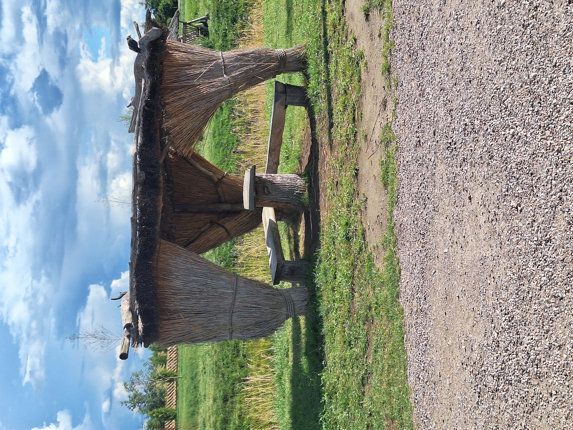 The image depicts an outdoor scene with a thatched gazebo constructed from straw and wood, featuring two rustic wooden benches and a central log table. The gazebo is situated in a grassy area adjacent to a path made of gravel and dirt. In the background, there is a body of water, lush green vegetation, and trees. The sky is partly cloudy with a mix of blue sky and white clouds. The overall setting suggests a natural, rural environment.