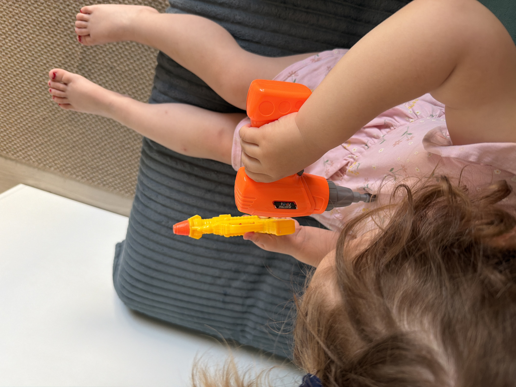 A toddler, possibly female, is lying on a gray cushion, holding a toy drill. The child has brown hair, fair skin, and is wearing a pink floral dress. The child is holding an orange and gray toy drill in her hands, with a yellow bit attached. The child's feet are visible at the top left of the image, with red nail polish on the toes. The background includes a white surface and a carpet. No information suggests this photo was taken in Pervomaysky, Russia.