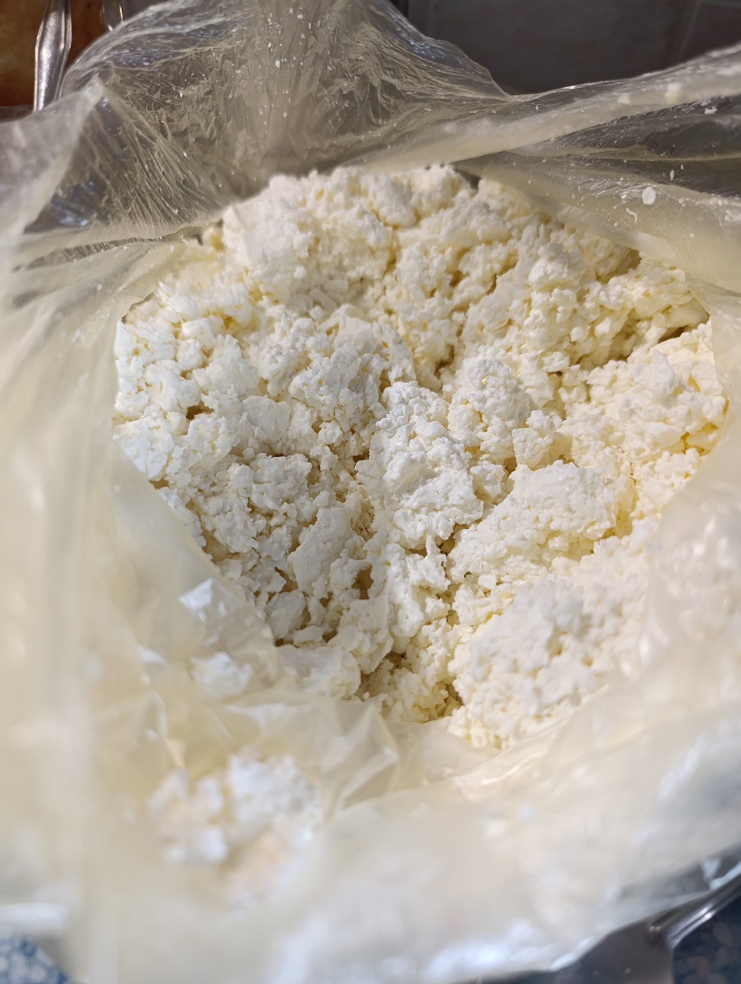 A close-up, overhead shot shows cottage cheese in a clear plastic bag. The bag is open at the top, revealing a white, lumpy texture of the cheese. The cheese fills most of the frame, with the plastic bag obscuring the edges. The background is blurred, but suggests an indoor setting.