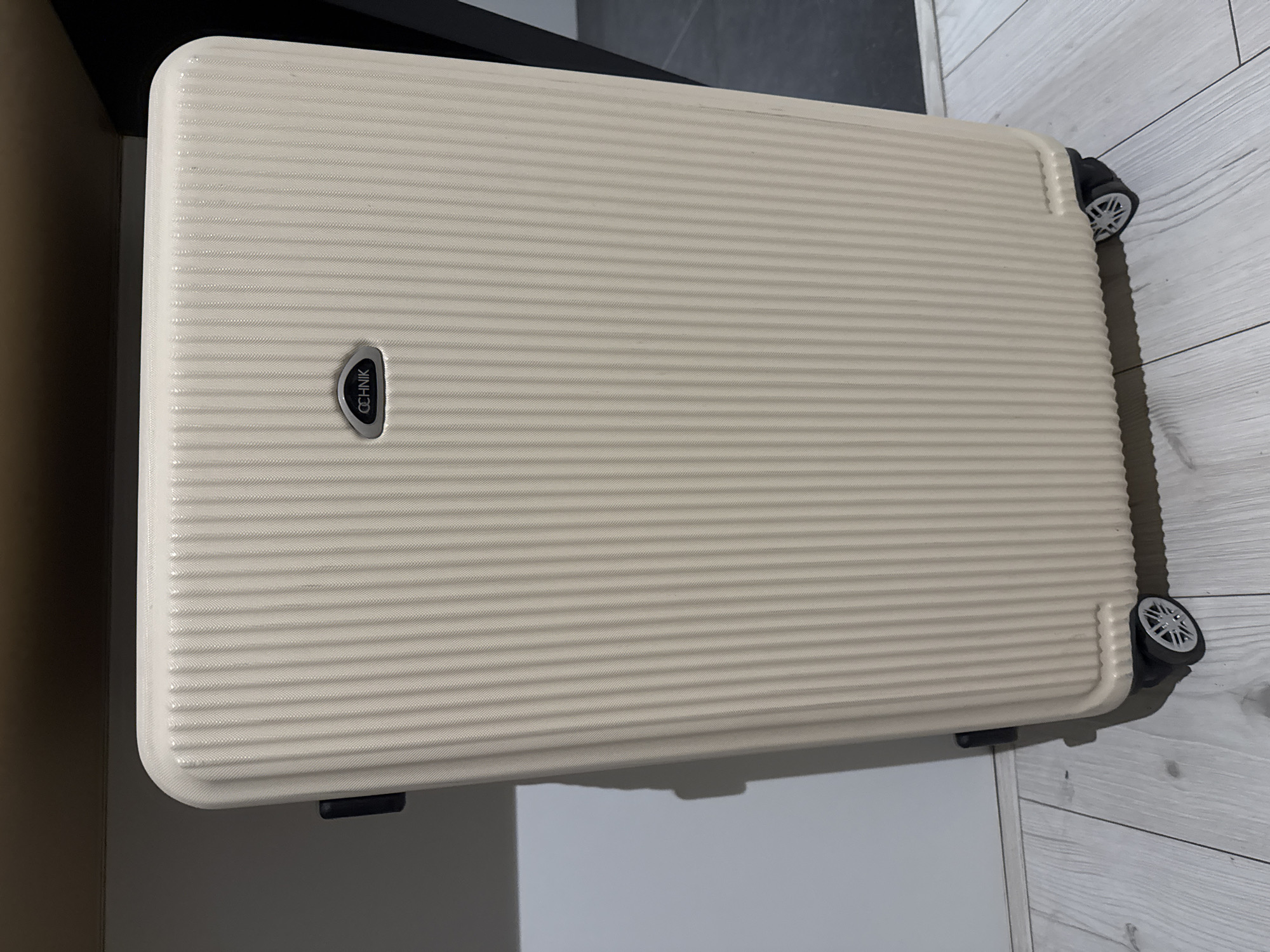 An off-white, ridged suitcase stands upright on a light-colored, wooden floor. The suitcase has a silver logo with the word "OCHNIK" on its front. It has black wheels at the bottom and a black handle on its left side. The suitcase is positioned against a white wall, with a dark brown surface above it. A shadow is cast on the wall behind the suitcase. The scene is indoors, in an unknown location.