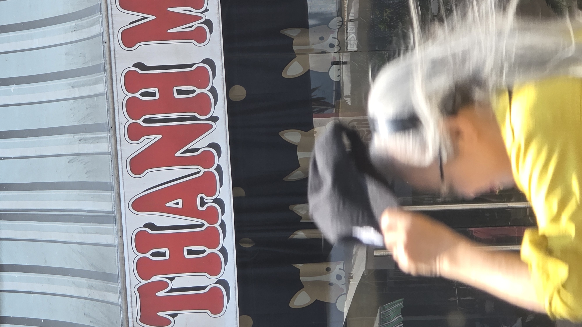 The scene shows a blurry image of a person wearing a yellow shirt and a cap, with a sign that reads "THANH NAM" in large red letters. The background includes a blue and white striped awning and a curtain with a dog pattern. This is Nha Trang, Vietnam, as the sign likely represents a local business name. The person seems to be in motion, possibly adjusting their cap.