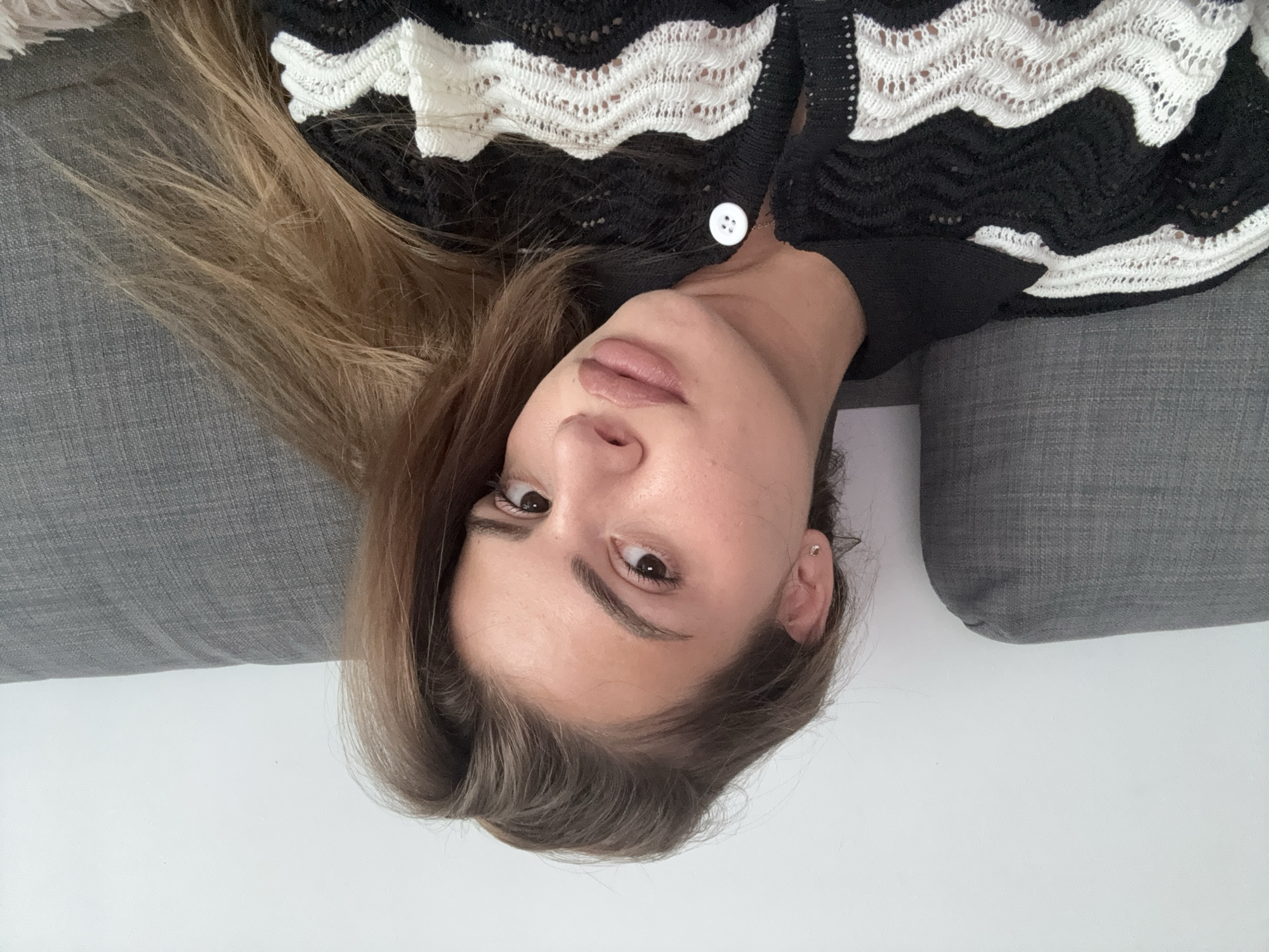 A medium close-up, high-angle shot captures a young woman lying on a gray upholstered surface, likely a sofa. The image is rotated, presenting her head upside down relative to a standard upright orientation. Her light brown hair is fanned out around her head and shoulders, blending into the gray fabric on one side and contrasting with a plain white background on the other.
She is wearing a black cardigan or top featuring prominent white, wavy, or zigzag knitted lace patterns across the chest and shoulders. A single white button fastens the garment near her collarbone.
Her face is centrally framed, with her dark eyes open and directed towards the camera, conveying a neutral yet direct expression. Her eyebrows are well-defined, and her full lips are painted with a matte nude or light brown lipstick. A small, subtle stud earring is visible in her right ear (viewer's left). The lighting is bright and even, suggesting an indoor setting. There is no visible text.