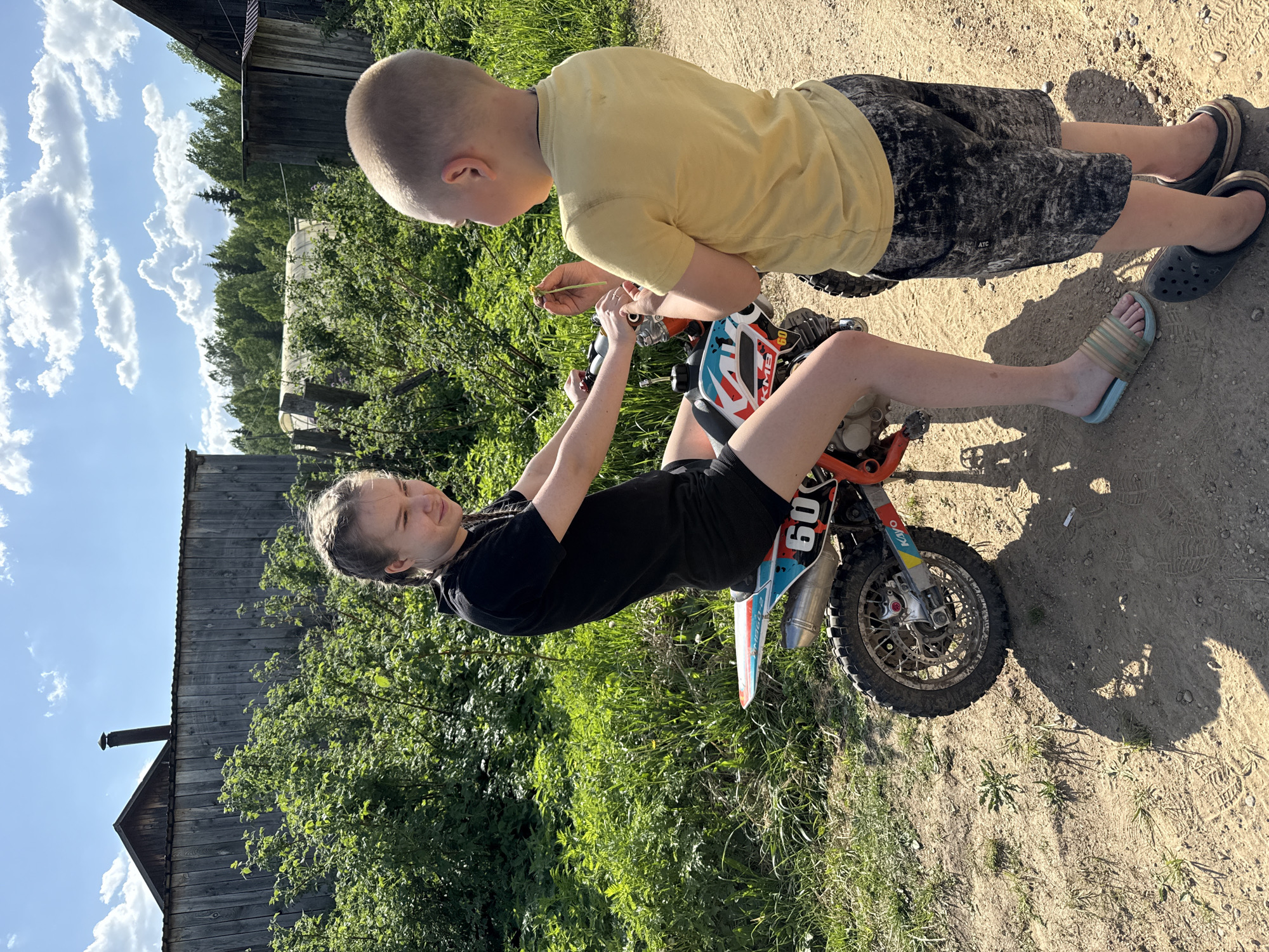 Two children are interacting with a small dirt bike in an outdoor setting. A girl with braided hair, wearing a black shirt and shorts, sits on the bike, holding the handlebars. A boy, wearing a yellow shirt and patterned shorts, stands next to her, holding a small twig and looking at the bike's controls. Both are wearing sandals. The bike has the number 60 on its side. The setting appears to be a dirt path next to a grassy area with vegetation. In the background are wooden buildings. Based on the query the location is Zhigalki, Russia.