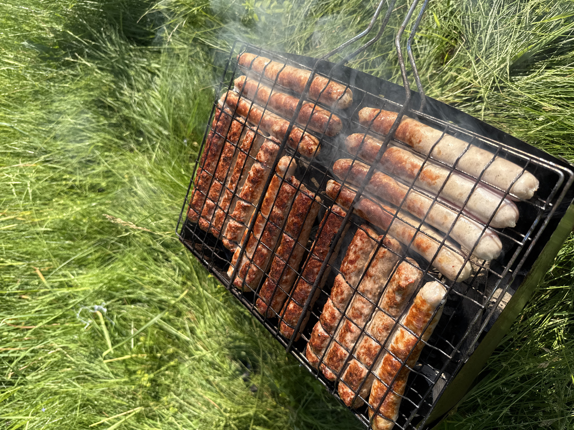 A close-up, high-angle shot captures sausages grilling on a metal rack placed on a barbecue. The sausages are arranged in rows, some appearing browned and cooked, while others are lighter in color. Wisps of smoke rise from the barbecue, mingling with the lush green grass that surrounds the scene. The grass is long and appears to be in a natural, uncultivated setting. The image is taken outdoors, suggesting a picnic or barbecue event. The location is identified as Korotsko, Russia, though no specific city landmarks or details confirm this. There are no people visible in the image.
