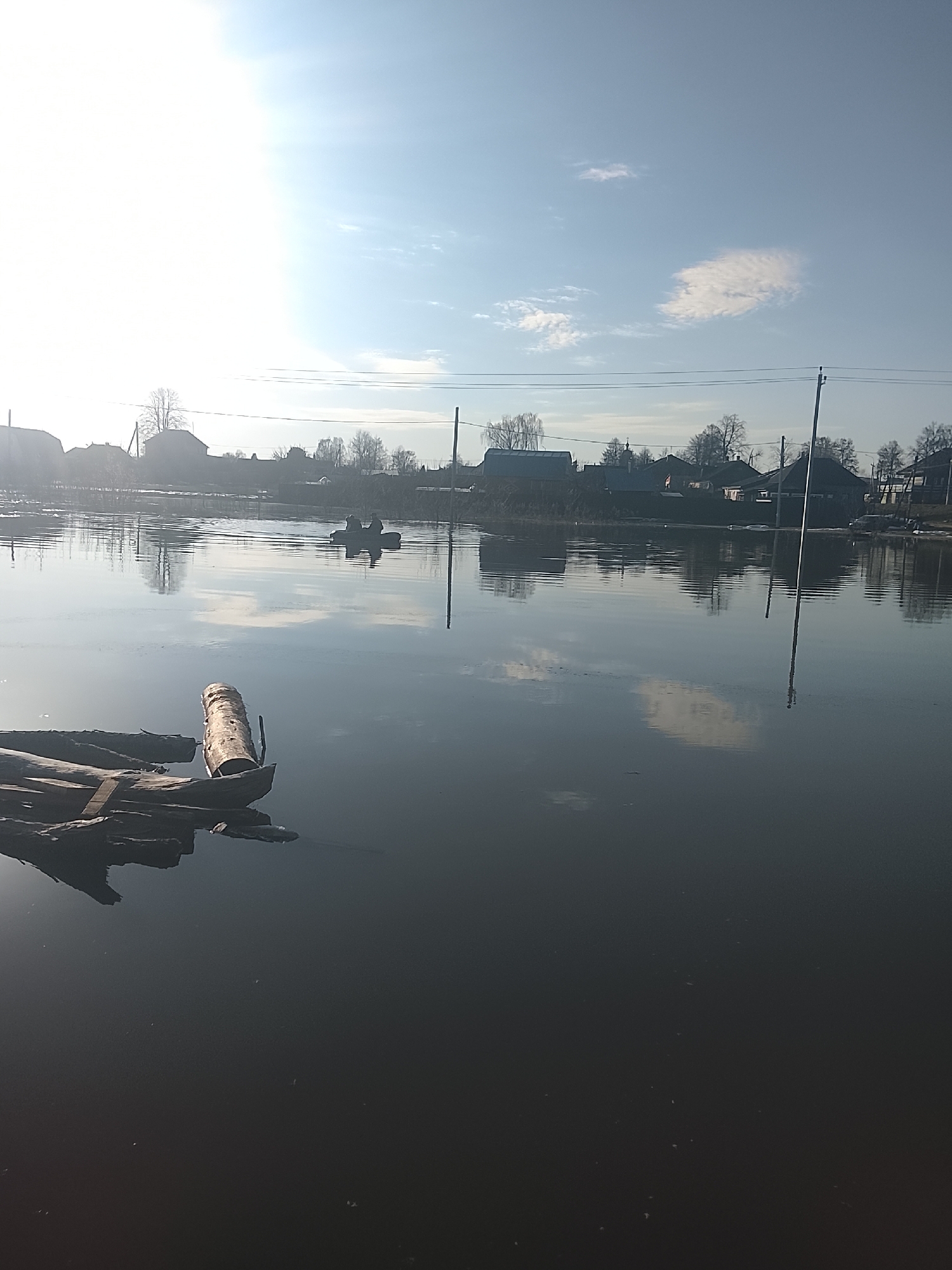 The image depicts a flooded area in Высокиничи, Russia. The water appears dark and still, reflecting the sky and surrounding buildings. In the foreground, a makeshift raft or grouping of logs is visible. Further in the distance, there is a small boat with two people. The background shows buildings and utility poles partially submerged in the water. The sky is light blue with scattered clouds. The overall impression is one of a flooded landscape within a residential area.