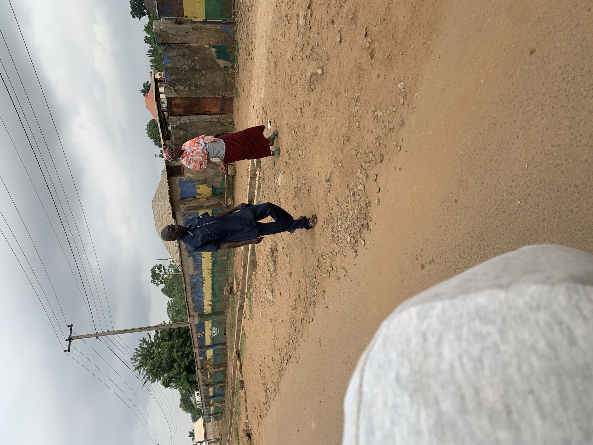 A dirt road in Maigizo, Nigeria, features a man walking parallel to the road and an older woman further along in a red skirt. Buildings line one side of the road; a power pole stands at the start. In the foreground, a large, blurred white area is seen on the right.