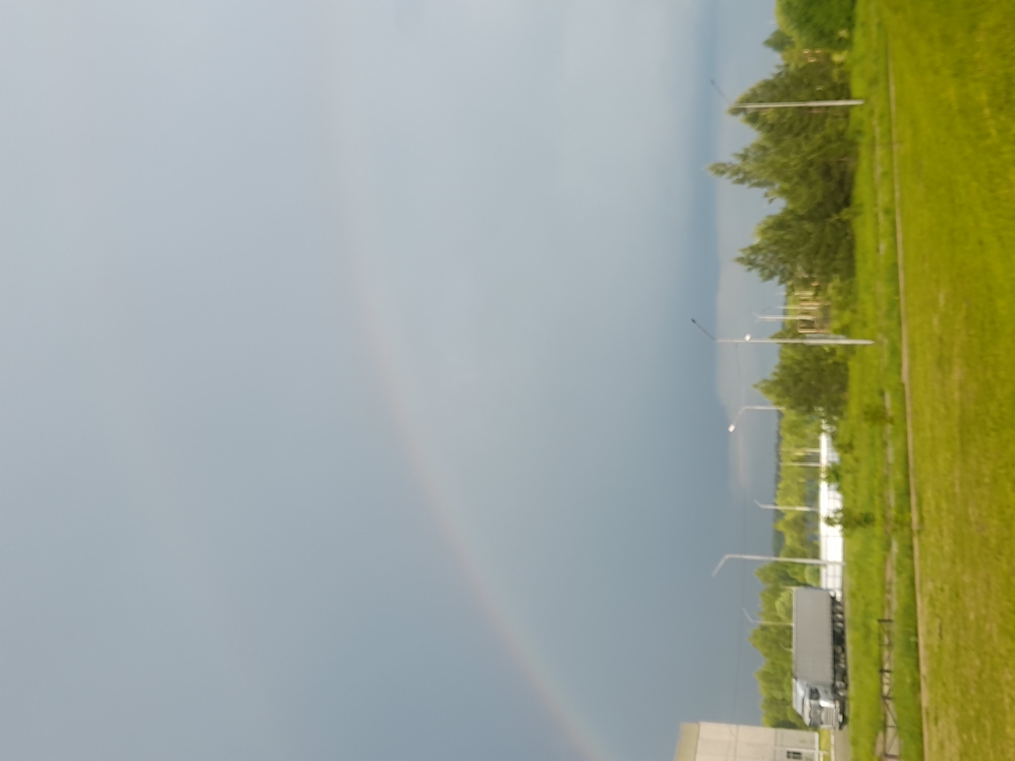 The image shows a landscape with a rainbow in the sky. The ground has green grass, trees, and light poles. A large truck is parked near a building, possibly in an industrial or commercial area. The sky is overcast, creating the conditions for the rainbow.