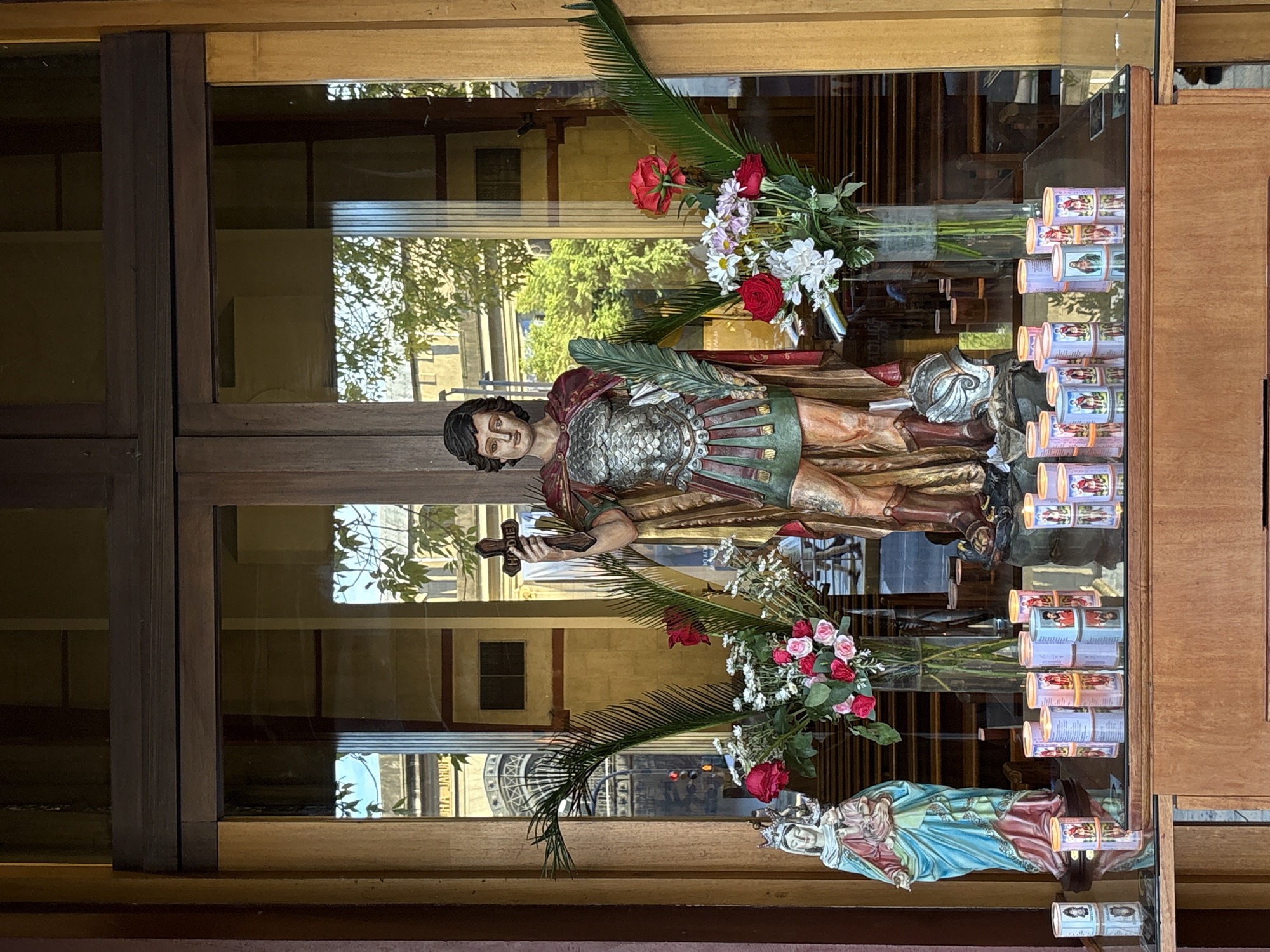 The image captures a vibrant religious devotional display, likely situated within a church or shrine in Buenos Aires, Argentina. The scene is viewed through a clear glass enclosure with a wooden frame.
At the heart of the display stands a statue of a male saint, depicted as a Roman soldier. He wears an intricately detailed silver cuirass over a green and red tunic with pteruges, and a flowing red cloak. In his left hand, he holds a small wooden cross, while a stylized helmet rests on the base beside his feet. His expression is solemn and serene.
To the right, partially visible, is a statue of the Virgin Mary, adorned with a crown and dressed in blue and pink robes, cradling the infant Jesus.
Both statues are surrounded by fresh floral arrangements in glass vases, featuring bouquets of red, pink, and white roses, white daisies, and dramatic green palm fronds, contributing to the ceremonial aesthetic.
To the right of the display, numerous cylindrical votive candles are neatly arranged in rows on a reflective surface. Many of these candles are lit, casting a warm glow, and each is adorned with colorful labels depicting various saints or religious figures.
The glass enclosure strongly reflects the external environment, revealing an outdoor urban setting. Visible reflections include lush green trees with abundant foliage, bright daylight, and multi-story buildings. A distinctive architectural feature resembling a dome or cupola is clearly reflected in the lower left pane of the glass. Faint, unreadable text can be observed on some candle labels and reflected building surfaces. The overall impression is one of pious reverence and spiritual offering.