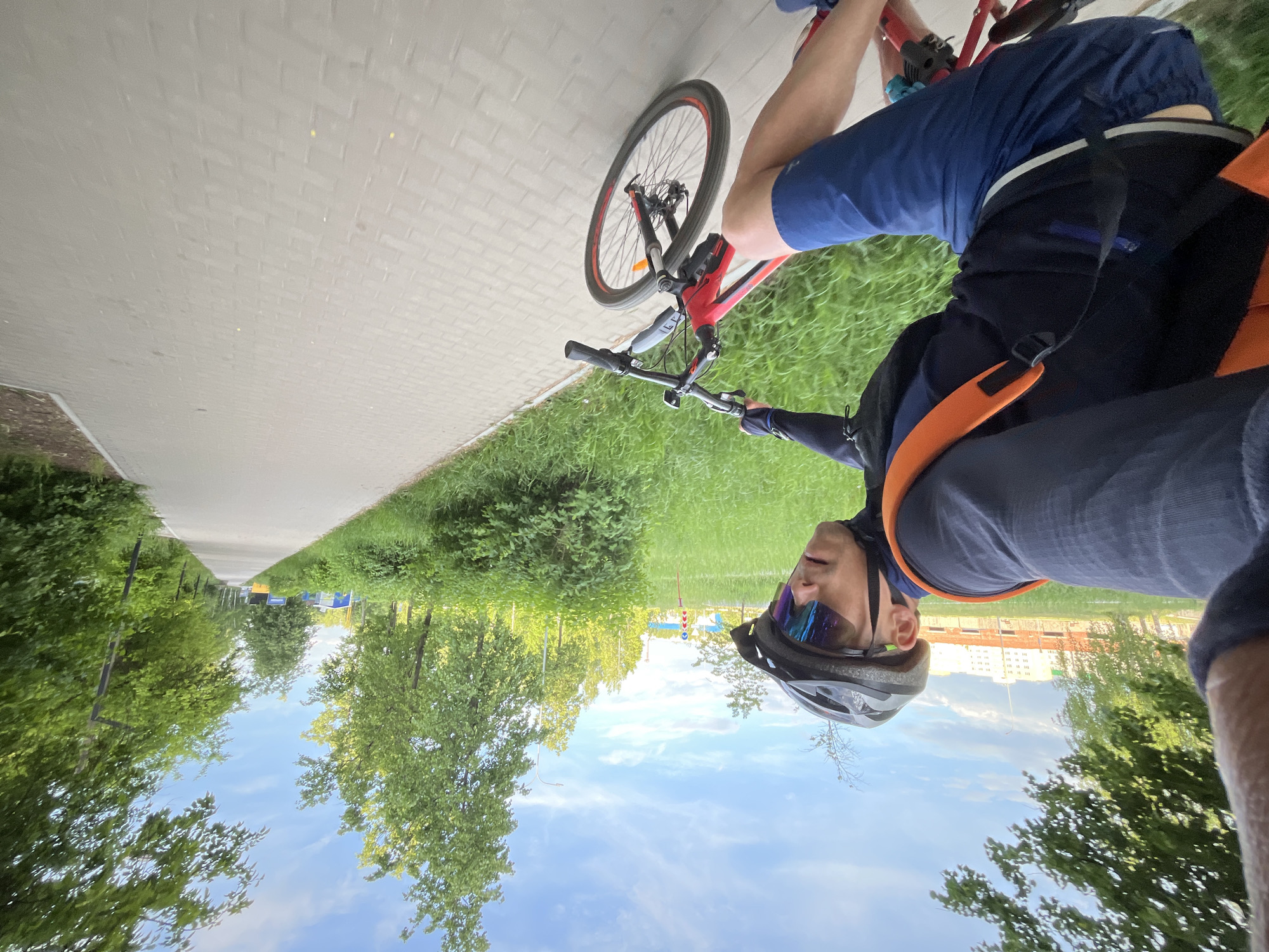 A man wearing a helmet and sunglasses is riding a red bicycle on a sidewalk in Chaikovsky, Russia. He is wearing blue clothing and an orange backpack. The sidewalk is made of light-colored paving stones and is bordered by green grass. Trees and blue sky are visible in the background. In the distance, a building is seen.