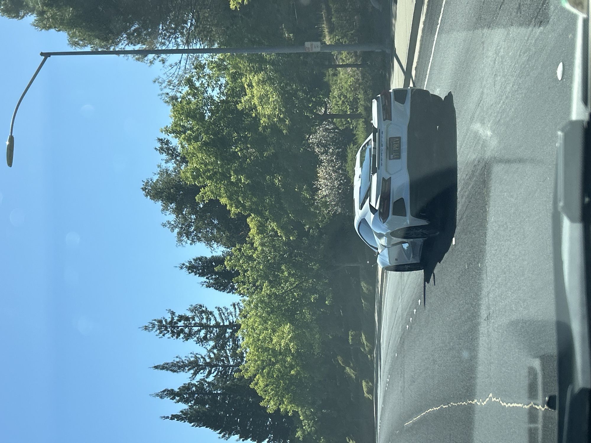 A white Chevrolet Corvette C8 is driving on a street in Roseville, California, USA. The car is traveling away from the camera on a multi-lane road. The car has a license plate that reads "YY PRIV". The road has clear lane markings and is bordered by trees and vegetation on the right side. A street light is visible on the right side of the image. The sky is clear and blue.