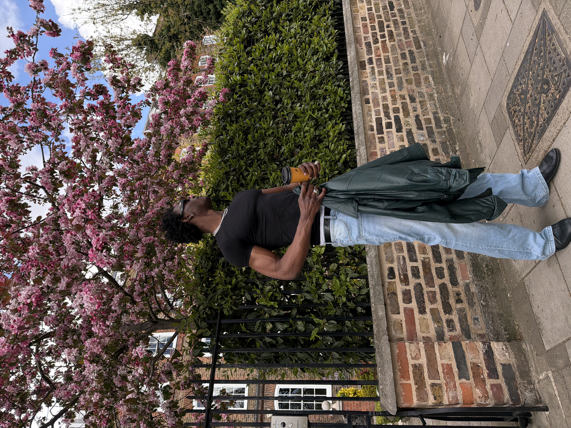 A man in sunglasses stands on a London street near flowering trees. He wears a black t-shirt, light blue jeans, and black shoes. He holds a brown cup in his right hand, and a dark green jacket is slung over his left shoulder. A brick wall borders the sidewalk where he stands. Behind him, a tall green hedge partially obscures a building with white-framed windows. A black metal gate stands in front of the building. The blossoming tree with pink flowers is on the left side of the frame.