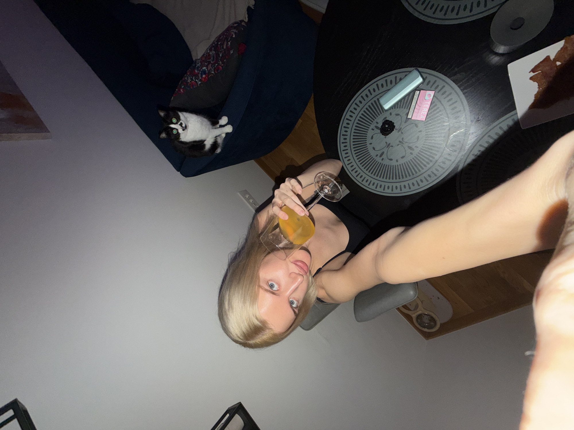 The image shows a woman with blonde hair and blue eyes taking a selfie while holding a glass of orange liquid, likely a cocktail, indoors. A tuxedo cat rests on a dark blue couch with a patterned pillow. Next to the couch is a round black table with a pack of cigarettes and lighter and a plate with a dark substance that may be food, such as brownies. The scene appears to be in a living room with a modern design and furniture. Based on the woman's previous posts and details visible in the image, the location is likely Belgrade, Serbia. The woman is holding the phone up at an angle to take the picture. A partial view of a chair is visible.