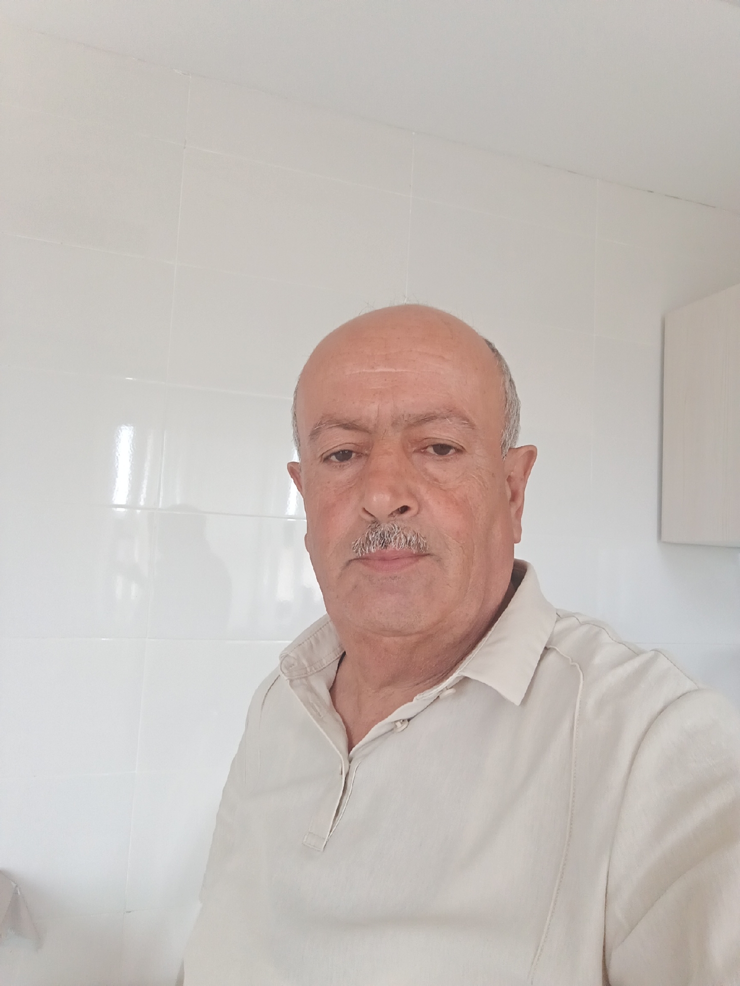 The image is a medium shot of an older man with a bald head and a mustache, wearing a light-colored collared shirt. He is looking directly at the camera. The background consists of white tiled walls, suggesting an indoor environment. There is a faint reflection on the tiles to the left of the man, and a portion of a white cabinet or wall unit is visible to the right. The lighting is bright and even, with no discernible shadows indicating the time of day or light source. The scene appears to be a domestic or utilitarian space, such as a bathroom or kitchen. The photograph is likely taken in Masazir, Azerbaijan, given the provided context.