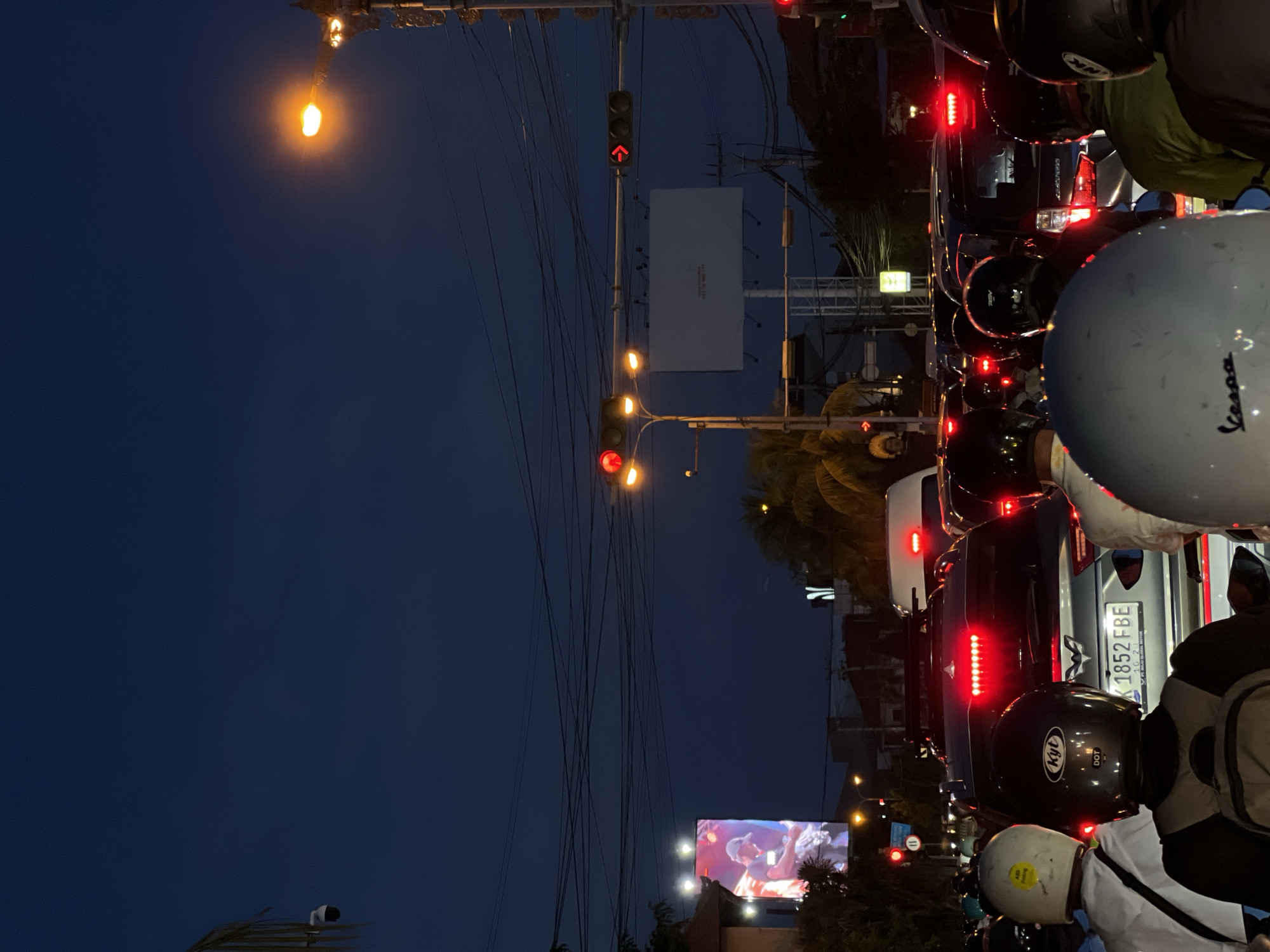 The image depicts a busy street scene at dusk or night in an urban environment, likely Denpasar, Indonesia. Heavy traffic is halted, with numerous motorcycles and cars stopped at what appears to be a red traffic light. The sky above is a deep navy blue, contrasting with the artificial lights illuminating the scene.
In the immediate foreground, the backs of several motorcyclists' helmets are prominent. Visible text includes "Vespa" on a white helmet, and "KYT DOT" on a black helmet. Another helmet has partially visible "INK" branding. People are seen wearing casual clothing, including a green shirt and a white shirt.
Further into the traffic, a dark SUV and a lighter-colored car are visible. The license plate "DK 1852 FBE" with "10-21" below is clearly legible on the lighter car, confirming the Indonesian context (DK is a common prefix for Bali vehicles). Red taillights and brake lights glow from the stopped vehicles.
Above the street, a complex network of overhead power lines crisscrosses the frame. Several streetlights emit an orange glow, and multiple traffic lights are visible; one displays a red arrow pointing upwards, while another shows a solid red circle, indicating a stop. Two billboards are present: a large, unlit white rectangular board, and a smaller, illuminated digital screen displaying an image of a person, likely an advertisement. A circular white sign with a red border and a black horizontal bar (indicating "no stopping" or "no parking") is also faintly visible on the left side of the road. The overall impression is a vibrant, congested city street after sunset.
