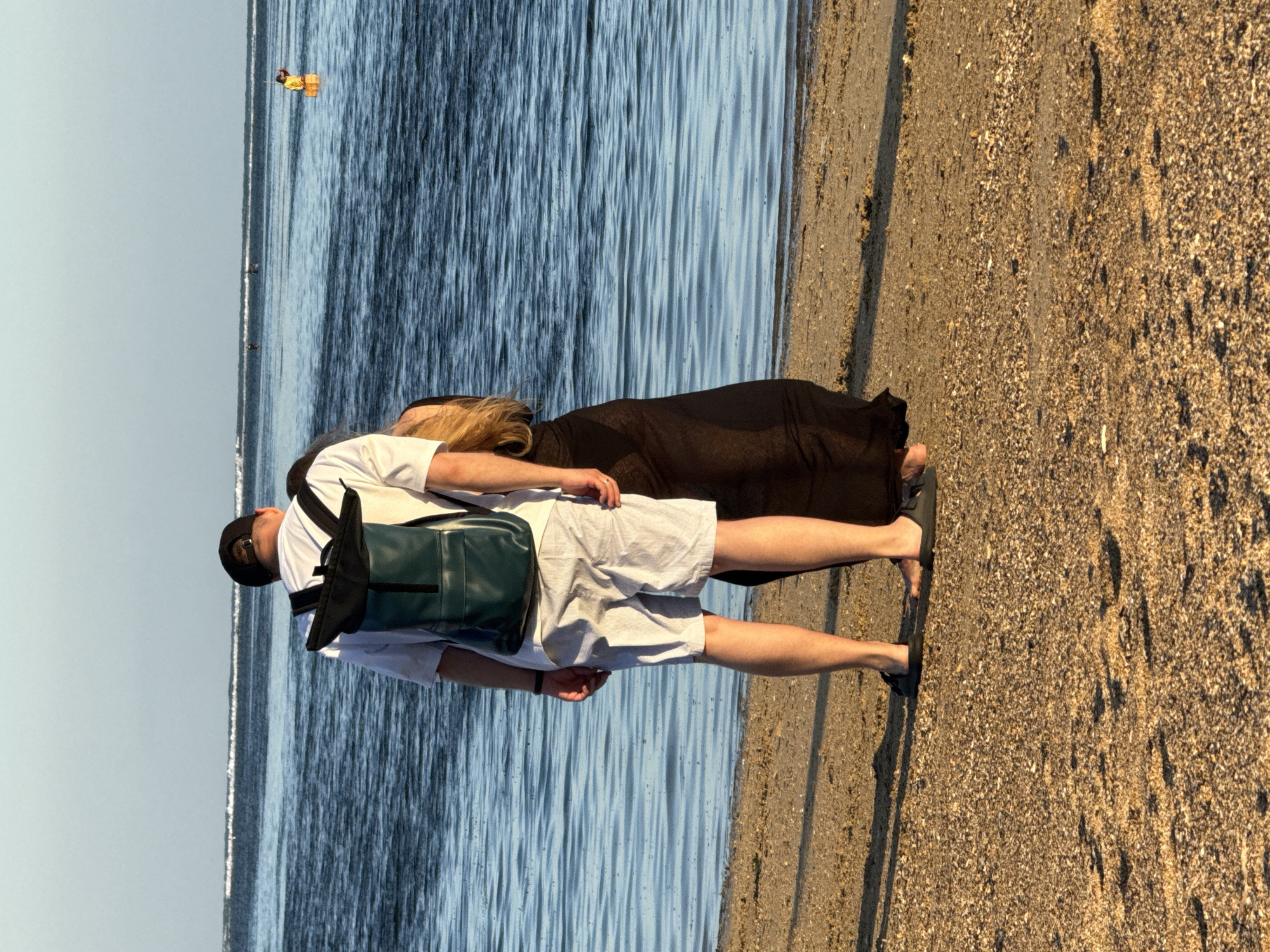 An unidentifiable man wearing a white t-shirt, light-colored striped shorts, and a dark baseball cap stands on a sandy beach facing the ocean, with a dark green backpack on his back. To his right, a woman in a long black dress stands partially obscured behind him, facing the same direction. The ocean is calm with gentle waves and a clear horizon. In the distance, two individuals are visible in the water. The scene is lit by natural daylight, suggesting it is daytime in Denpasar, Indonesia, likely at a beach location such as Sanur Beach or Seminyak Beach. The ground is covered in sand and small pebbles.