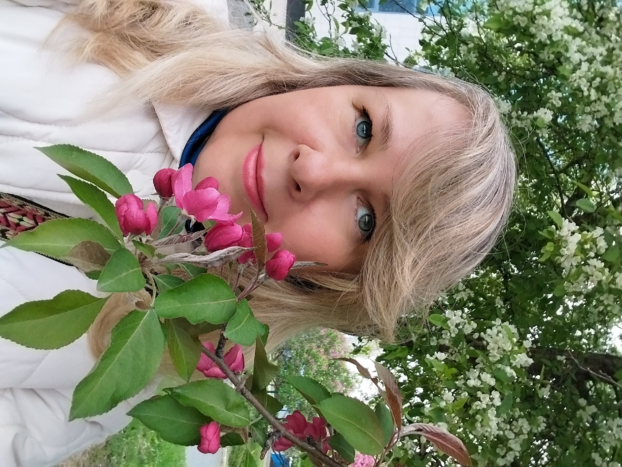 A selfie of a fair-skinned woman with blonde hair and blue eyes, taken outdoors near blooming trees. She holds a branch with pink flowers in the foreground. The background includes trees with white blossoms. The location is Orenburg, Russia.