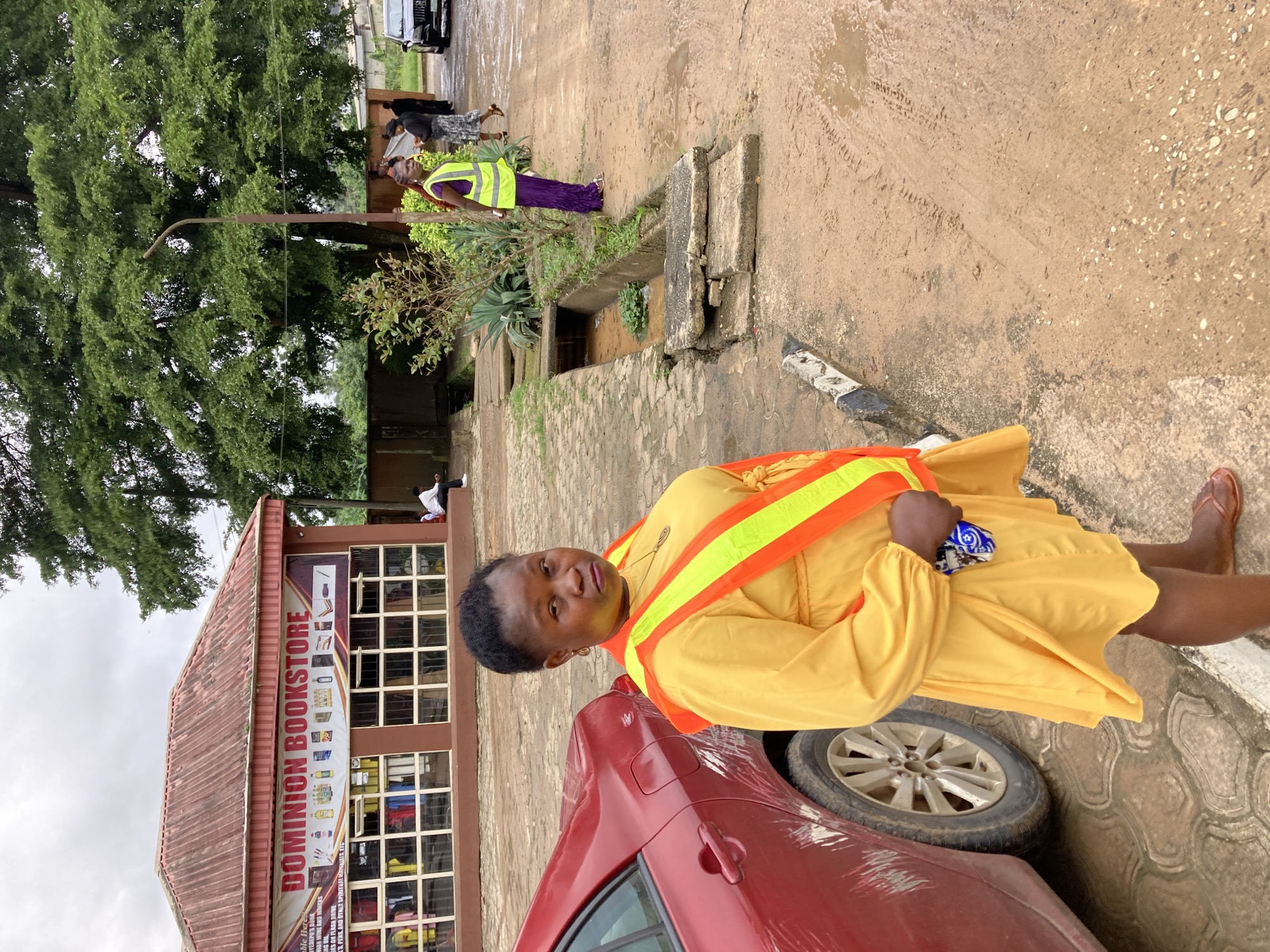 A medium shot captures a woman standing in front of a red car and a bookstore in Aba, Nigeria. The woman, positioned in the foreground, wears a bright yellow dress and an orange and yellow reflective vest. She is looking directly at the camera with a neutral expression. The red car is to her left, and the Dominion Bookstore, with a banner displaying its name and various products, is in the background. Several other people are visible in the middle ground and background, some near the bookstore and others walking. The environment appears to be an outdoor setting with a paved area, a drainage ditch, and lush greenery from trees. The sky is overcast, suggesting a cloudy day.