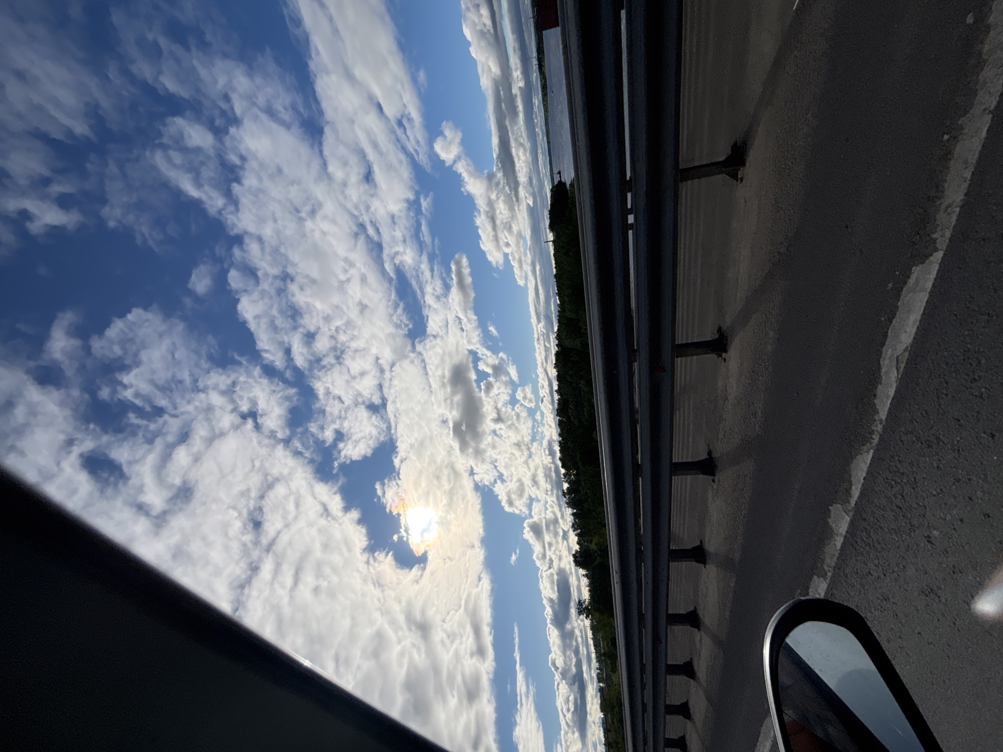 The image depicts a view from a vehicle, likely a car, on a bridge or elevated roadway. To the right is a gray asphalt road surface with a metal guardrail along the edge. Beyond the guardrail is a line of trees and then water, possibly a river or lake. To the left, a blue sky with scattered white clouds is visible. The sun is obscured by some of the clouds. Part of the car's interior (the window frame) and a side-view mirror are visible. The image appears to be taken in motion. It is impossible to confirm the location is Yaroslavl, Russia, with the information provided.