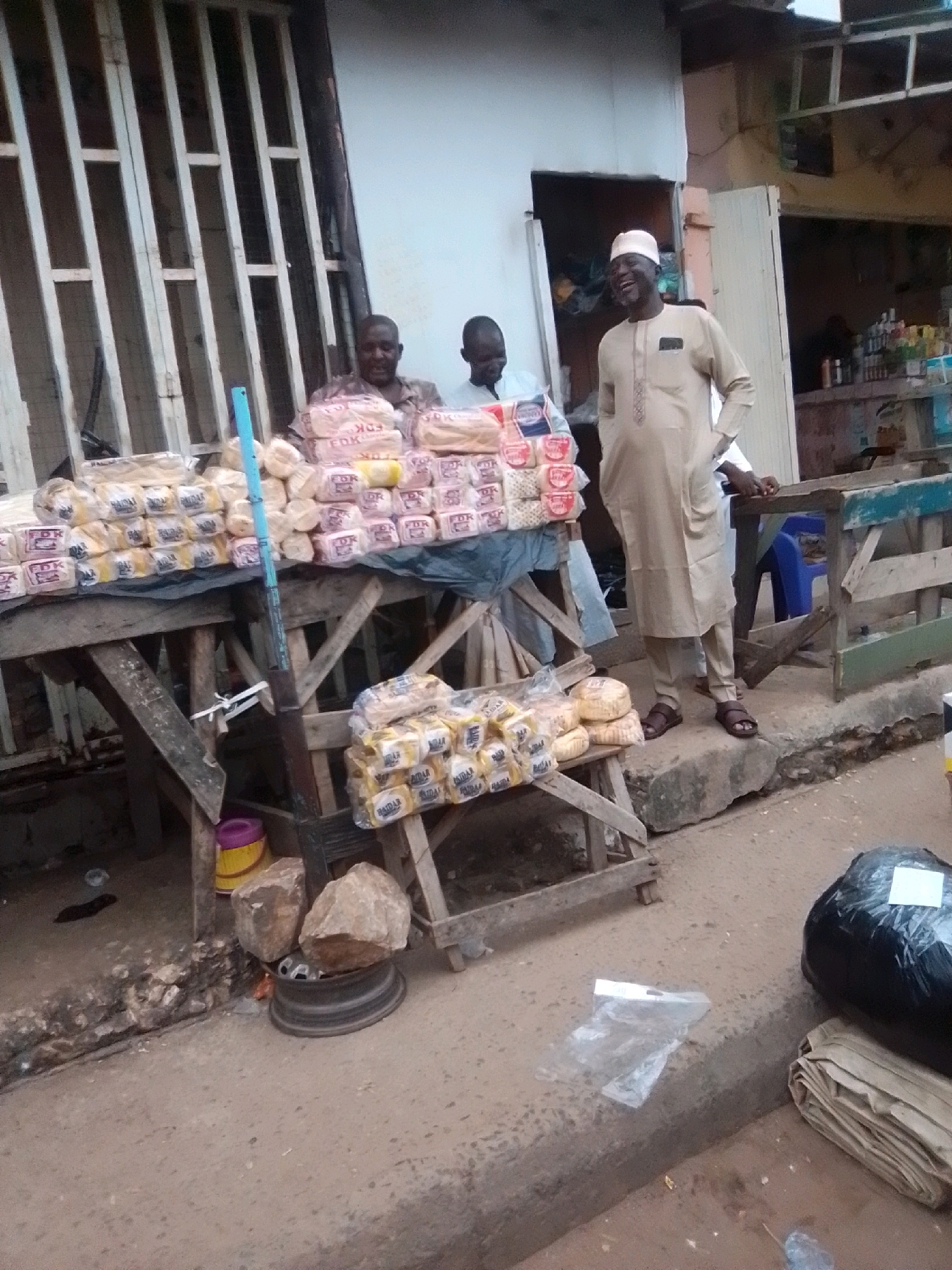 In Arawa, Nigeria, a street market scene unfolds. Three men are present, two behind a stall laden with packaged bread, and one standing in front of it. The man in front, wearing a light-colored traditional garment and a cap, is laughing. The stall is constructed from rough wood and displays stacks of bread wrapped in plastic. To the right of the stall, another man in a light-colored shirt stands next to a wooden counter. The ground is paved with concrete and dirt, with some debris scattered around. In the foreground, a large black plastic bag and a stack of folded tan material are visible on the ground. The overall environment suggests a bustling outdoor marketplace.