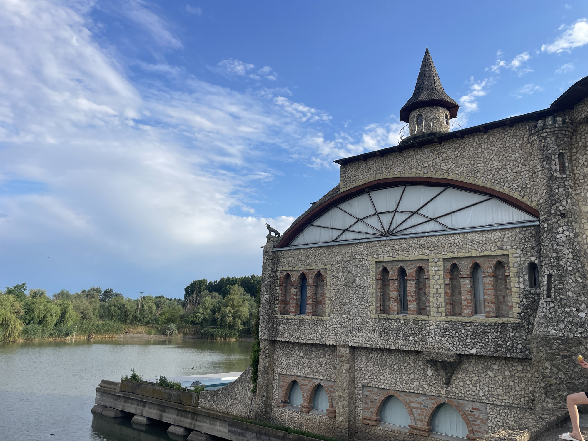 A multi-story stone building with a conical tower stands on the bank of a body of water. The building's facade is constructed from rough-hewn stones of varying sizes and colors. The structure features several arched windows on the lower levels and a large arched window with a segmented glass design on the upper level. A small, dark statue of an animal, possibly a wolf or dog, is perched on the roofline above the large window. The conical tower has a dark, shingled roof and a small, round window. The building is situated in a natural environment with lush green trees and reeds lining the opposite bank of the water. The sky above is a vibrant blue with scattered white clouds. A person is partially visible on the right side of the image, standing on a stone ledge and holding a yellow object. The scene is set in Colibași, Moldova.