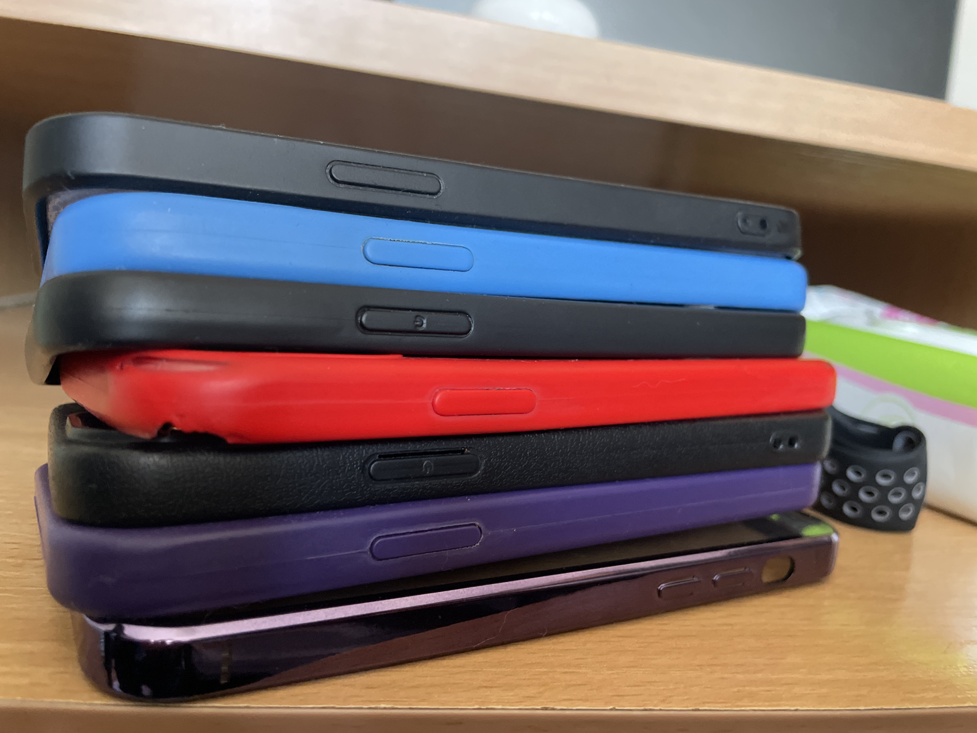 A stack of seven cell phone cases is on a table. From top to bottom, the cases are black, blue, black, red, black, purple, and dark purple. A black object with holes is next to the cases on the right. The background consists of a light-colored surface and some blurred items. The photo was taken indoors. The location is unknown, but presumably this is not the city of Asenovgrad, Bulgaria, as no evidence points towards it.
