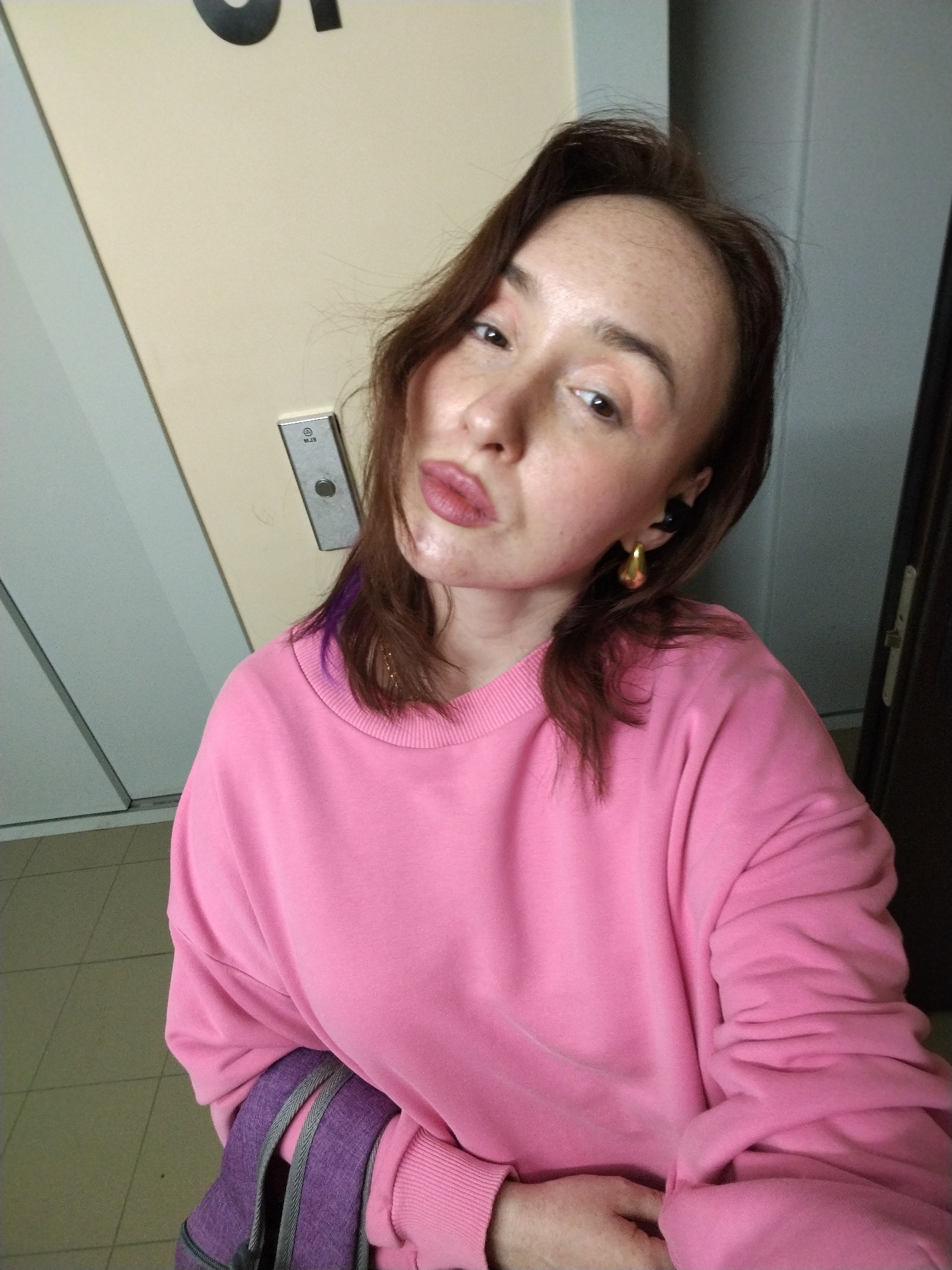 A young woman is captured in a close-up selfie indoors, looking directly at the camera with a slight pout. She has light skin with visible freckles, dark brown hair with a distinct purple streak on her left side, and wears light pink eyeshadow and lip gloss. Her accessories include a black in-ear earbud and a gold-colored, teardrop-shaped earring in her right ear, along with a thin gold chain necklace. She is dressed in a bright pink, long-sleeved crewneck sweatshirt. A purple backpack with grey straps is partially visible in the foreground, likely resting on her lap.
The setting appears to be an indoor hallway or common area in a building in Izhevsk, Russia. Behind her, a light beige wall features a silver elevator call button panel with a single dark circular button. Faint text, possibly "WJB" or "MJB," along with a small circular logo, can be seen on this panel. A large, dark, partially cropped letter, resembling an 'O' or 'C', is visible on the wall at the upper left. The floor is covered with light-colored square tiles. A light-colored door, possibly an elevator door, is reflected on the left, while a darker door or wall section is on the right. The lighting suggests an ambient indoor environment.