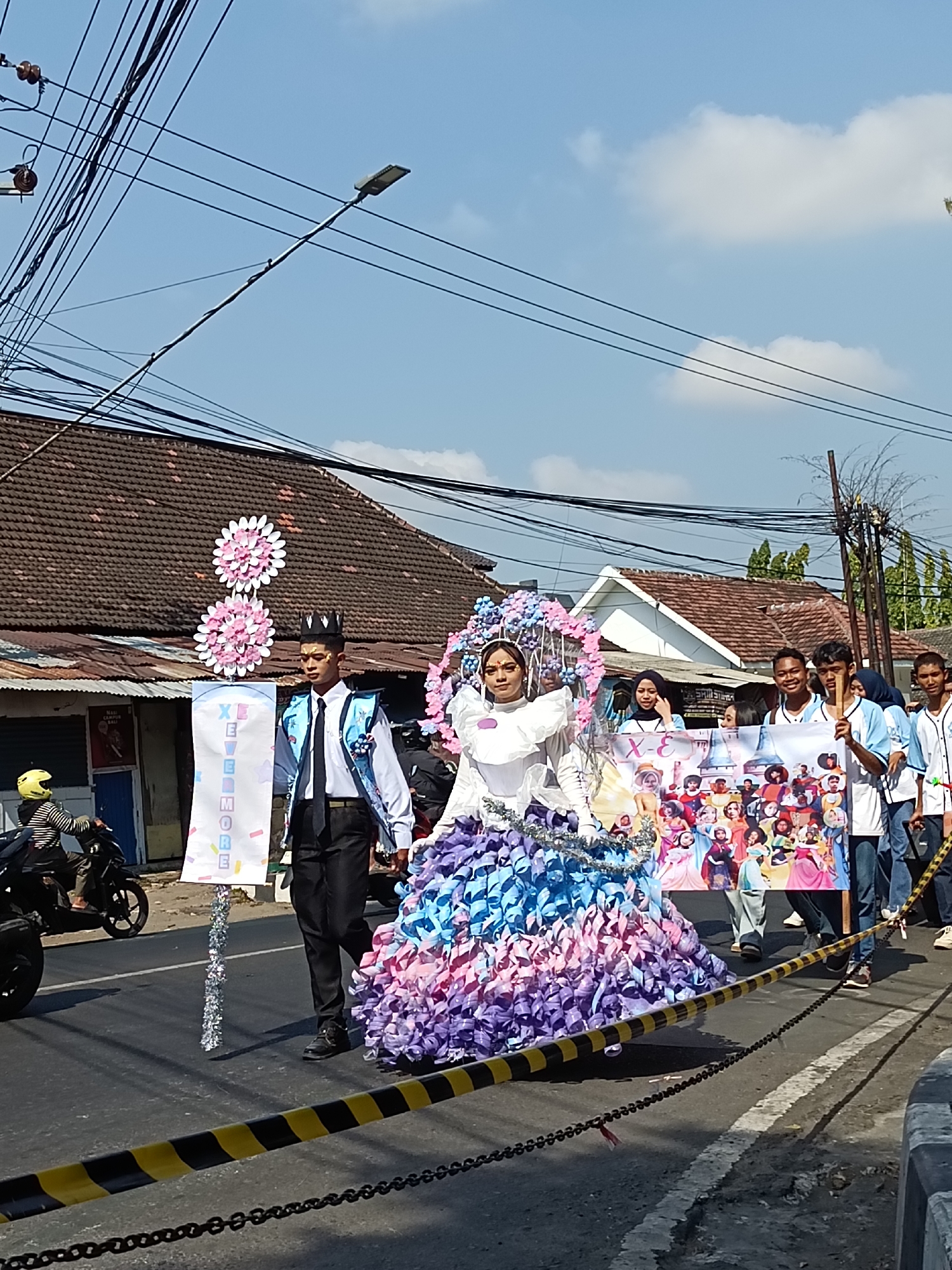 In Kediri City, Indonesia, a parade is taking place on a street lined with buildings and power lines. The main subjects are two individuals in elaborate costumes. One, appearing to be a man, wears a dark crown, a white shirt, a dark tie, and a patterned vest, standing next to a banner with pink floral decorations. Beside him is a person in a very large, colorful dress made of purple, pink, and blue ribbon-like material, topped with a large, elaborate headdress. Behind them, a group of people walk, some holding a large banner featuring cartoon princesses. In the background, a motorcycle rider with a yellow helmet is visible, and several other people are observing the parade. The scene is captured on a bright, sunny day.