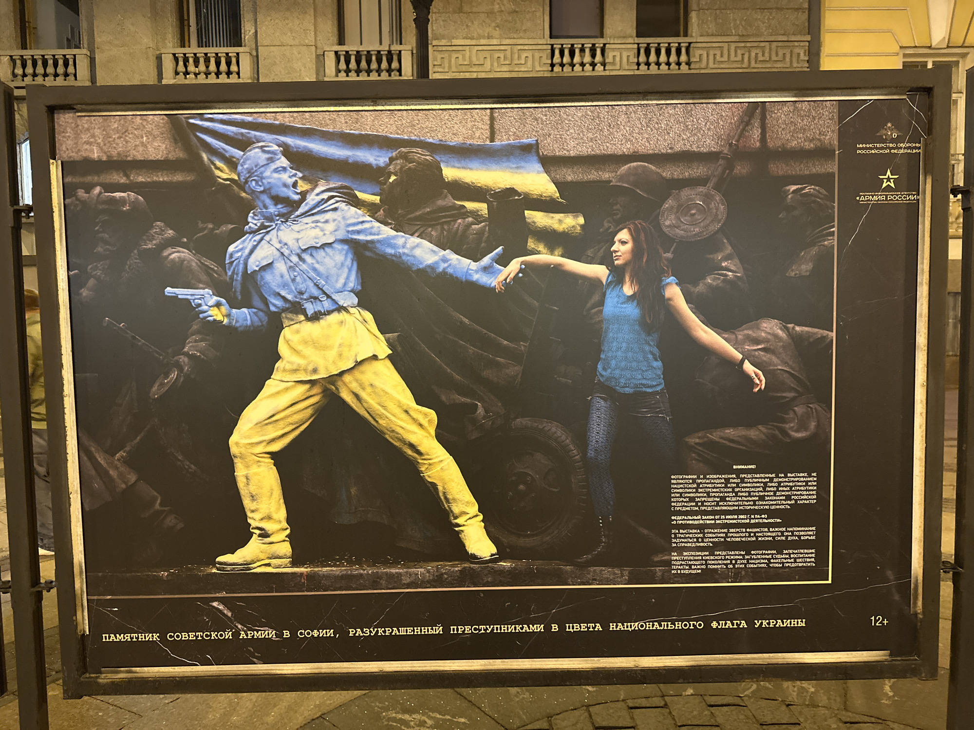 The image shows a framed poster, located in Saint Petersburg, Russia. The poster depicts a photograph of the Soviet Army monument in Sofia, Bulgaria, which has been painted in the colors of the Ukrainian flag. The painting shows the blue and yellow of the flag applied to the figures of the soldiers. A woman stands in front of the figures, extending her hand toward one of them. The top right of the poster displays the logo of the Ministry of Defense of the Russian Federation and the logo of "Army Russia" in the same region of the poster. Text is written below the photo, which reads "A monument to the Soviet Army in Sofia, decorated by criminals in the colors of the national flag of Ukraine". Smaller text outlines that images do not promote any extremist symbols, and are for informational purposes. At the bottom-right of the photo is a "12+" label.