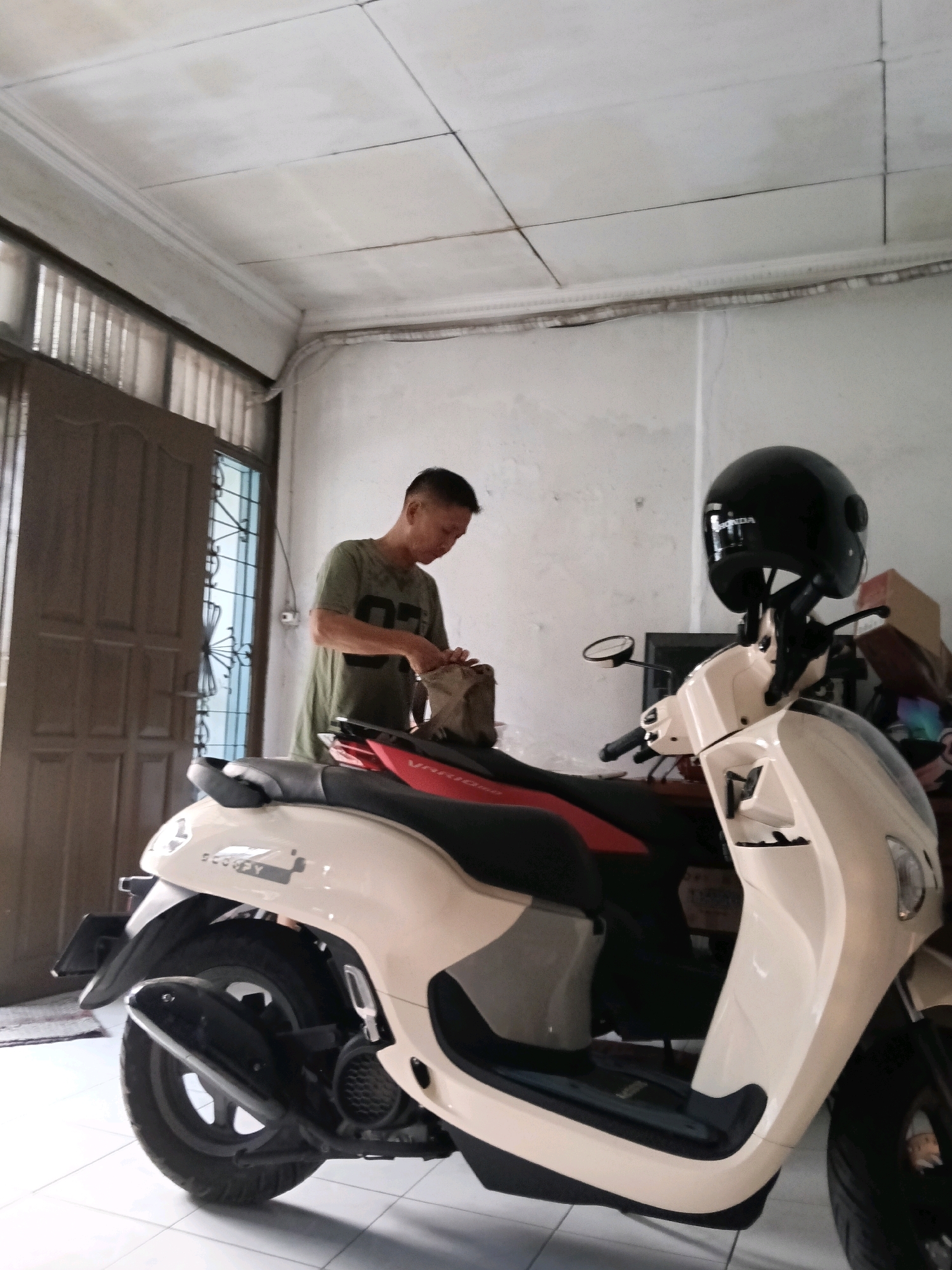 A man in a green t-shirt is standing in front of a cream-colored scooter, reaching into a bag on the seat. He is wearing a black helmet, which is placed on the handlebars of the scooter. The scooter is parked indoors, possibly in a garage or residential area, with a tiled floor and white walls. A brown door is visible to the left of the man. The ceiling is white with visible paneling and trim. The scene appears to be located in Jatimulya, Indonesia.
