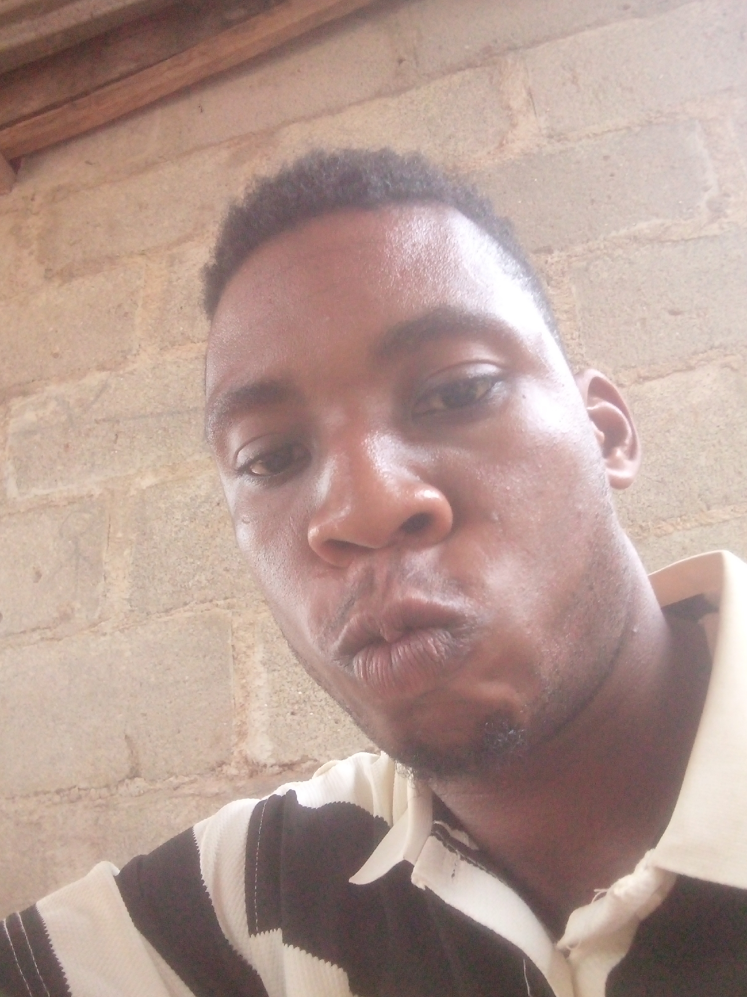 A close-up selfie of a dark-skinned man with short, dark hair. He is wearing a black and white striped shirt with a white collar. He is making a kissing face. The background is a wall made of light brown, rectangular blocks and some wooden boards. The location appears to be indoors. The city and area are Unknown, Unknown.