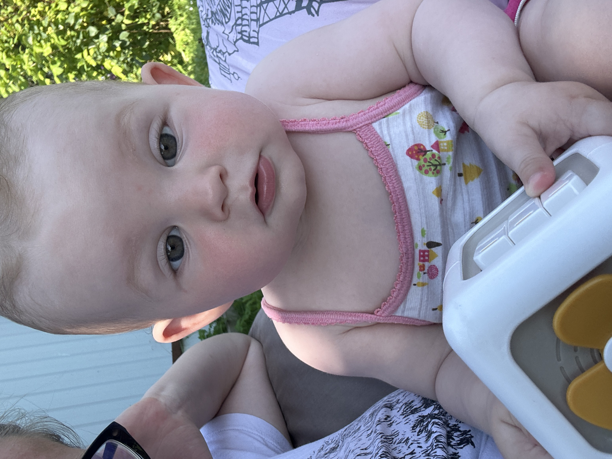 A young infant is seated outdoors, facing the camera. The infant has light skin, rosy cheeks, and dark grey eyes. They are wearing a white tank top with a pink trim and a pattern of houses and trees. The infant is holding a white and yellow toy in their hands. In the background, there is greenery and a light-colored structure, possibly a fence or building. A person wearing glasses is partially visible on the left side of the frame. The scene appears to be in a residential outdoor setting in Kalitsino, Russia.